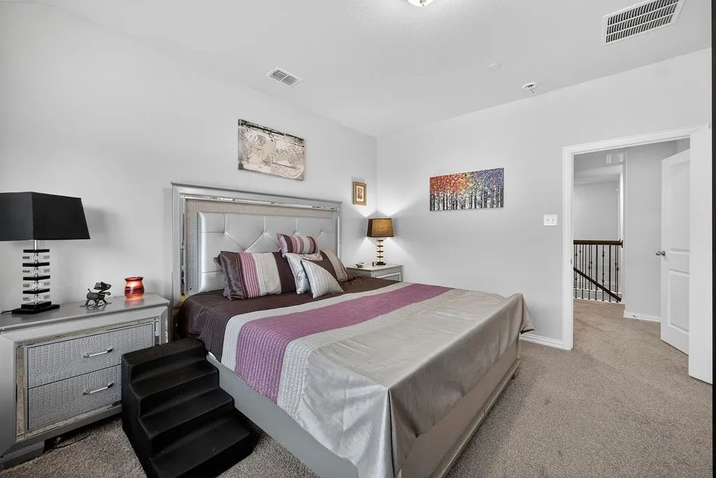 Carpeted bedroom featuring a decorative upholstered headboard, white walls, and a doorway opening to a hallway with stair access