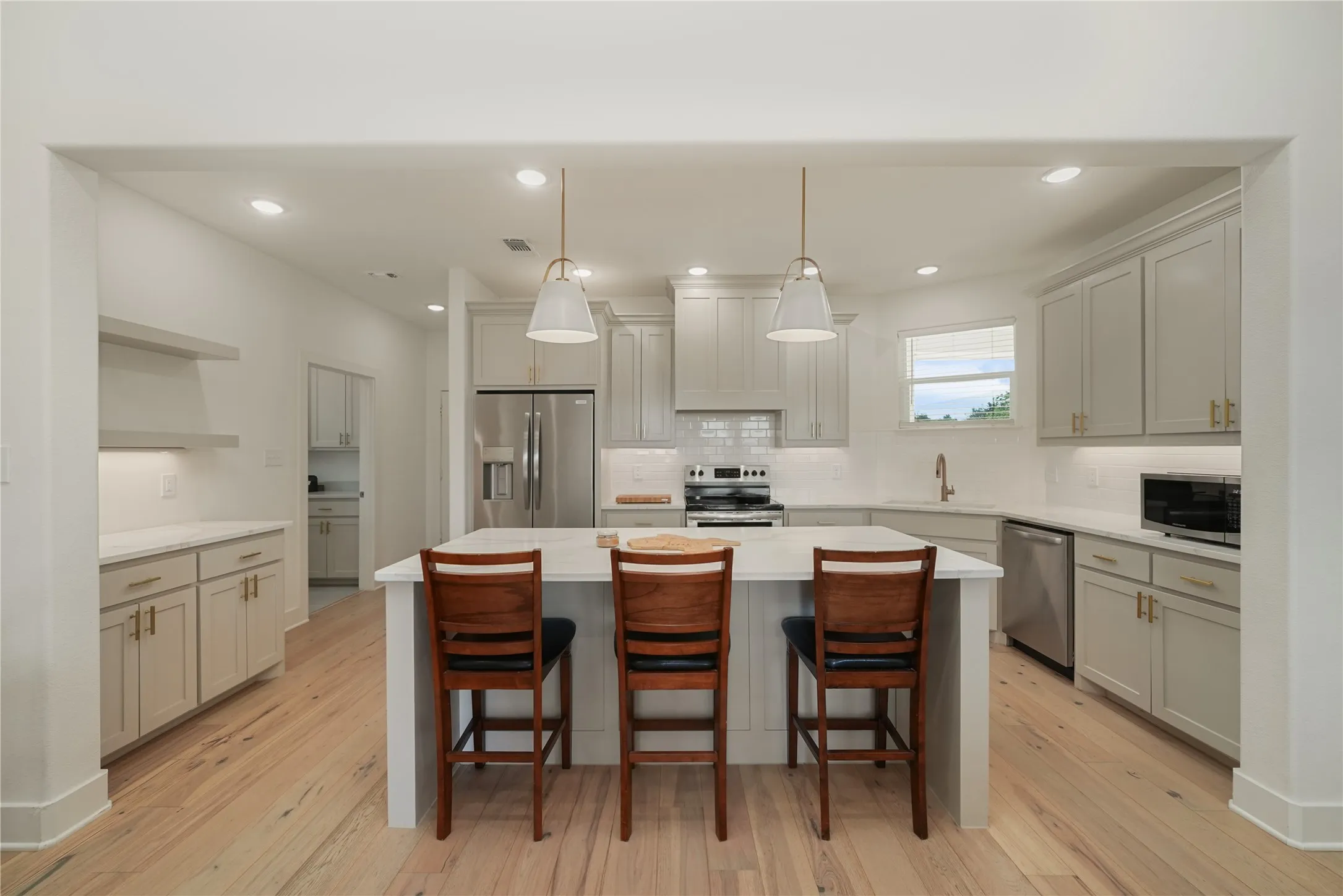 Modern kitchen featuring a central island with seating, light wood-finish flooring, white countertops, gray cabinetry with gold-tone hardware, and stainless steel appliances
