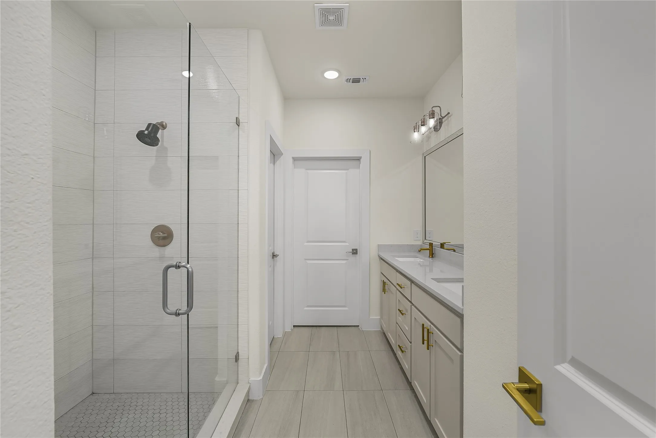 Spacious bathroom featuring a glass-enclosed shower with subway tile surround and hexagonal tile floor, dual vanity with undermount sinks and quartz countertop, brushed gold fixtures, and wood-finish tile flooring