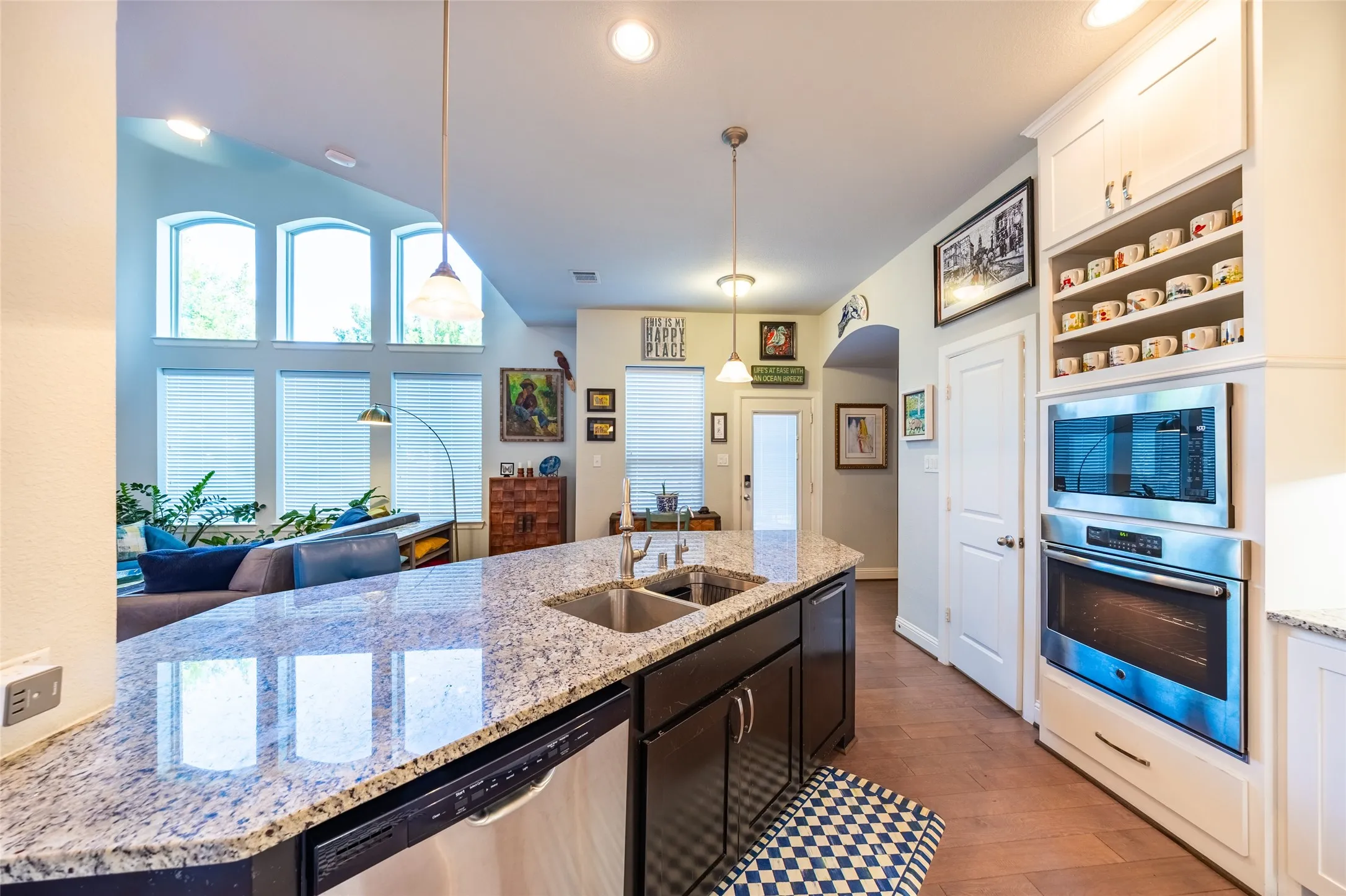 Open-concept living area featuring a kitchen island with dual sinks and granite-finish countertop, stainless steel dishwasher, and built-in wall oven and microwave