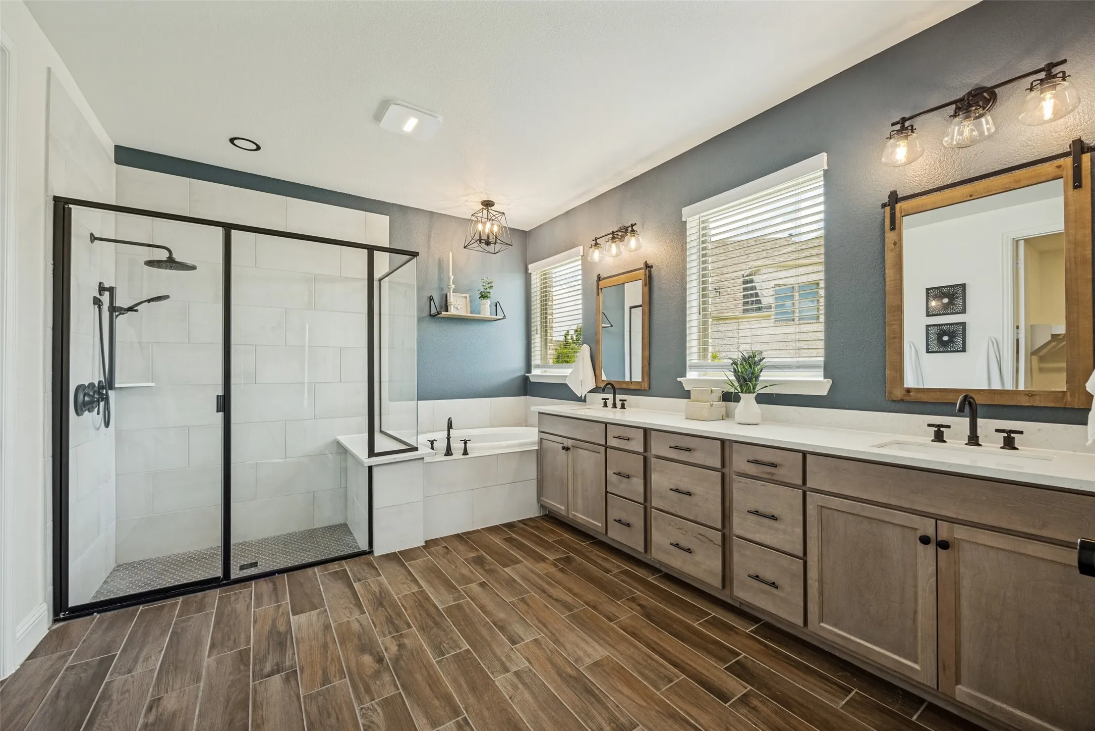 Spacious bathroom featuring a double vanity with white countertops, wood-finish flooring, a standalone tub, and a shower with black-framed glass enclosure