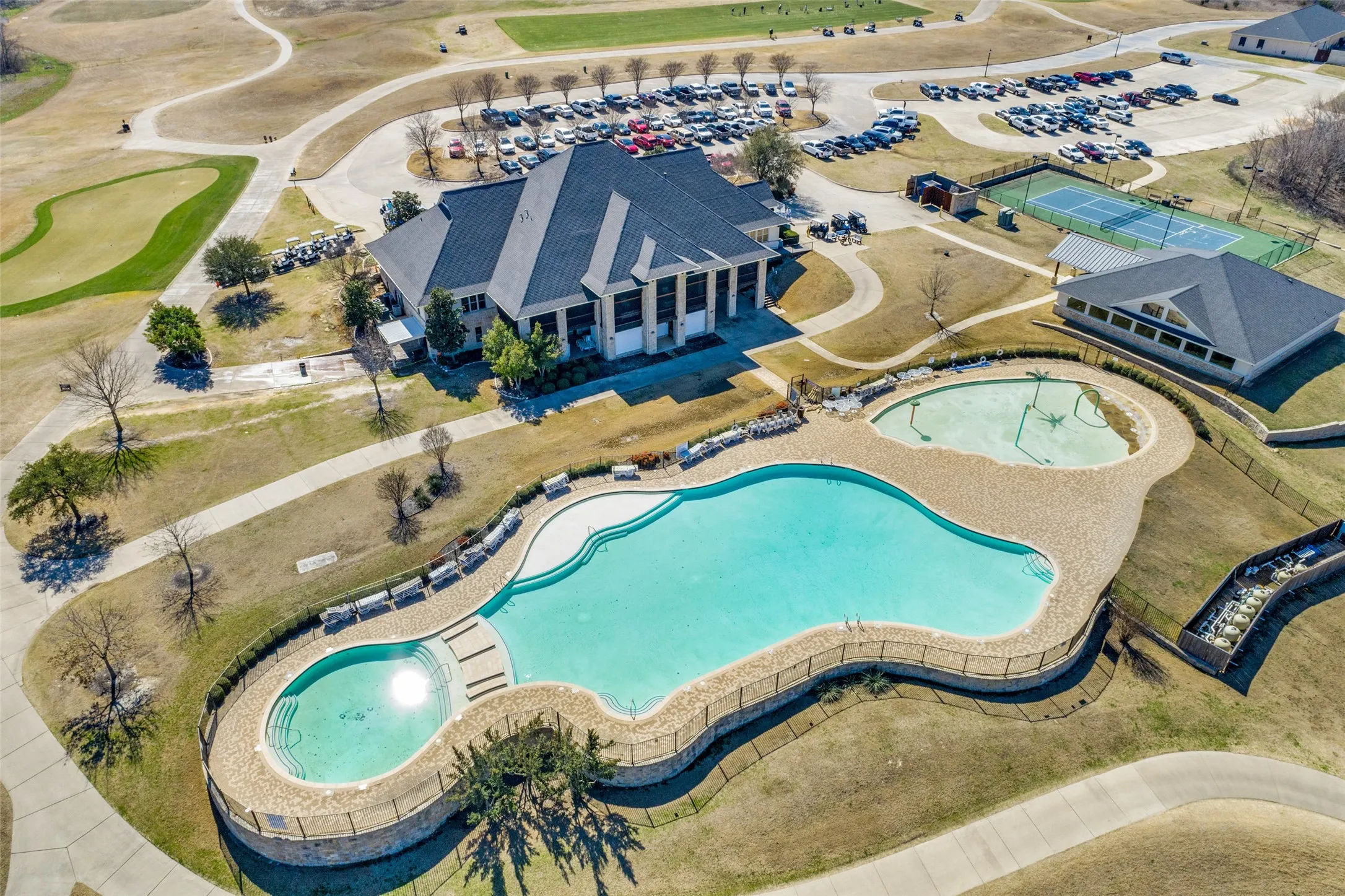 Expansive outdoor amenity area featuring a large swimming pool, a smaller adjacent pool with water features, tennis courts, and a spacious clubhouse building.