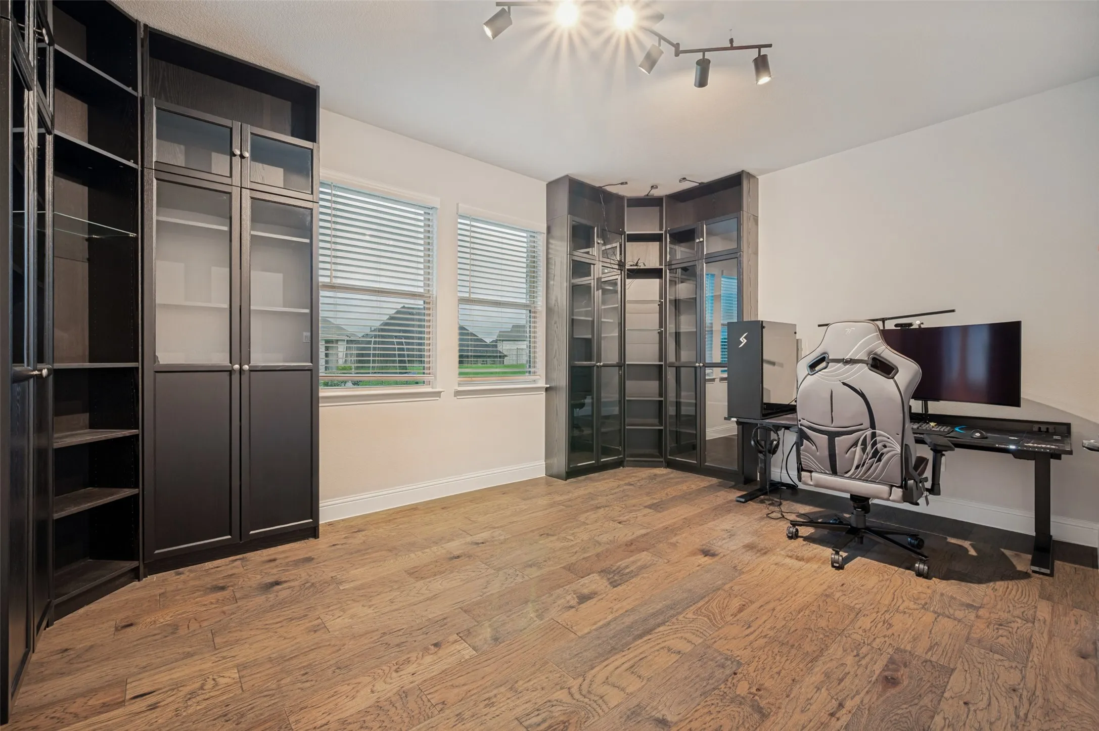 Versatile room featuring extensive built-in dark cabinetry with glass panel doors, wood-finish flooring, and two windows with horizontal blinds.