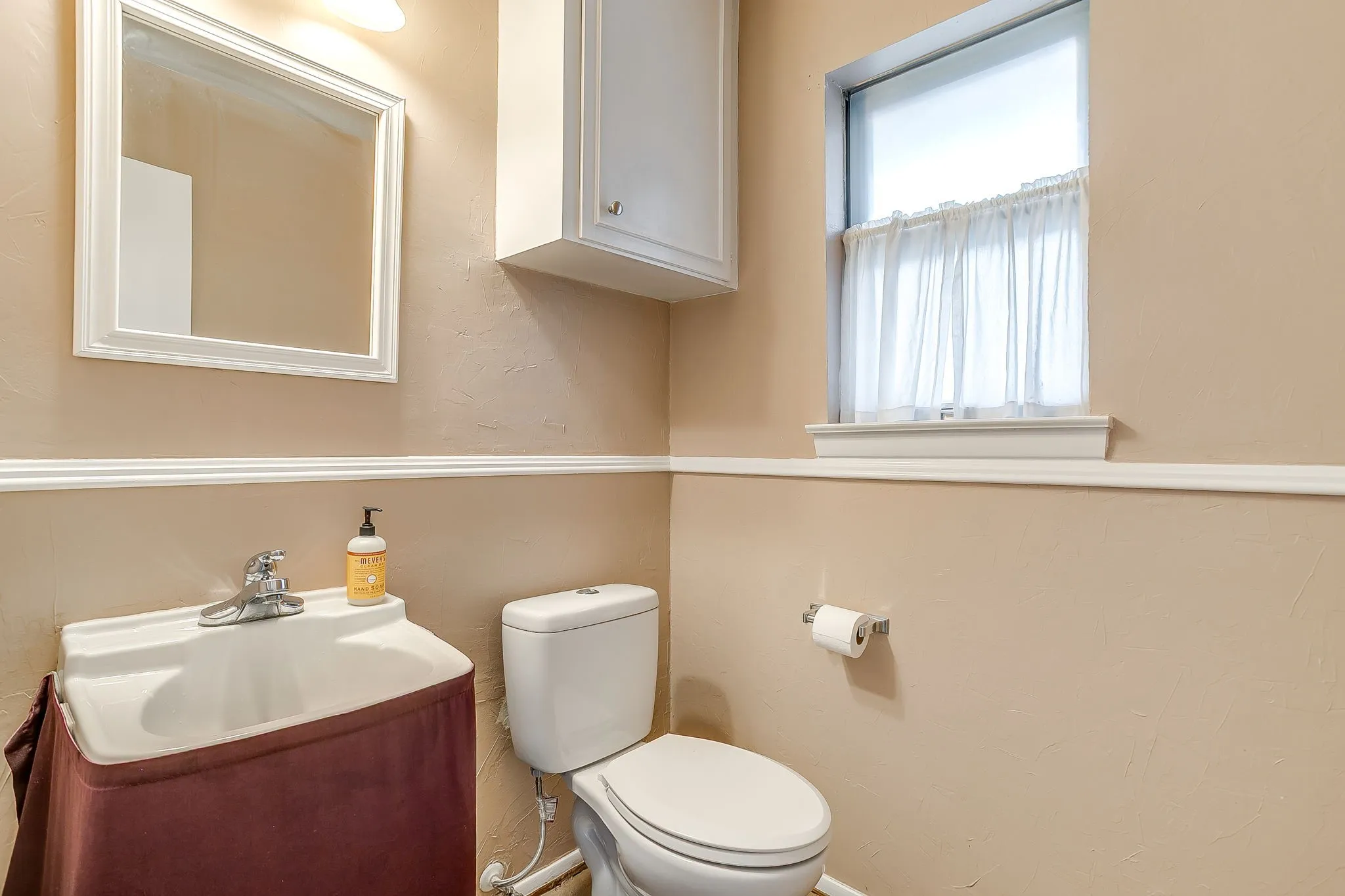 Half-bathroom with white framed mirror, overhead lighting, white wall-mounted cabinet, and a window with a sheer curtain