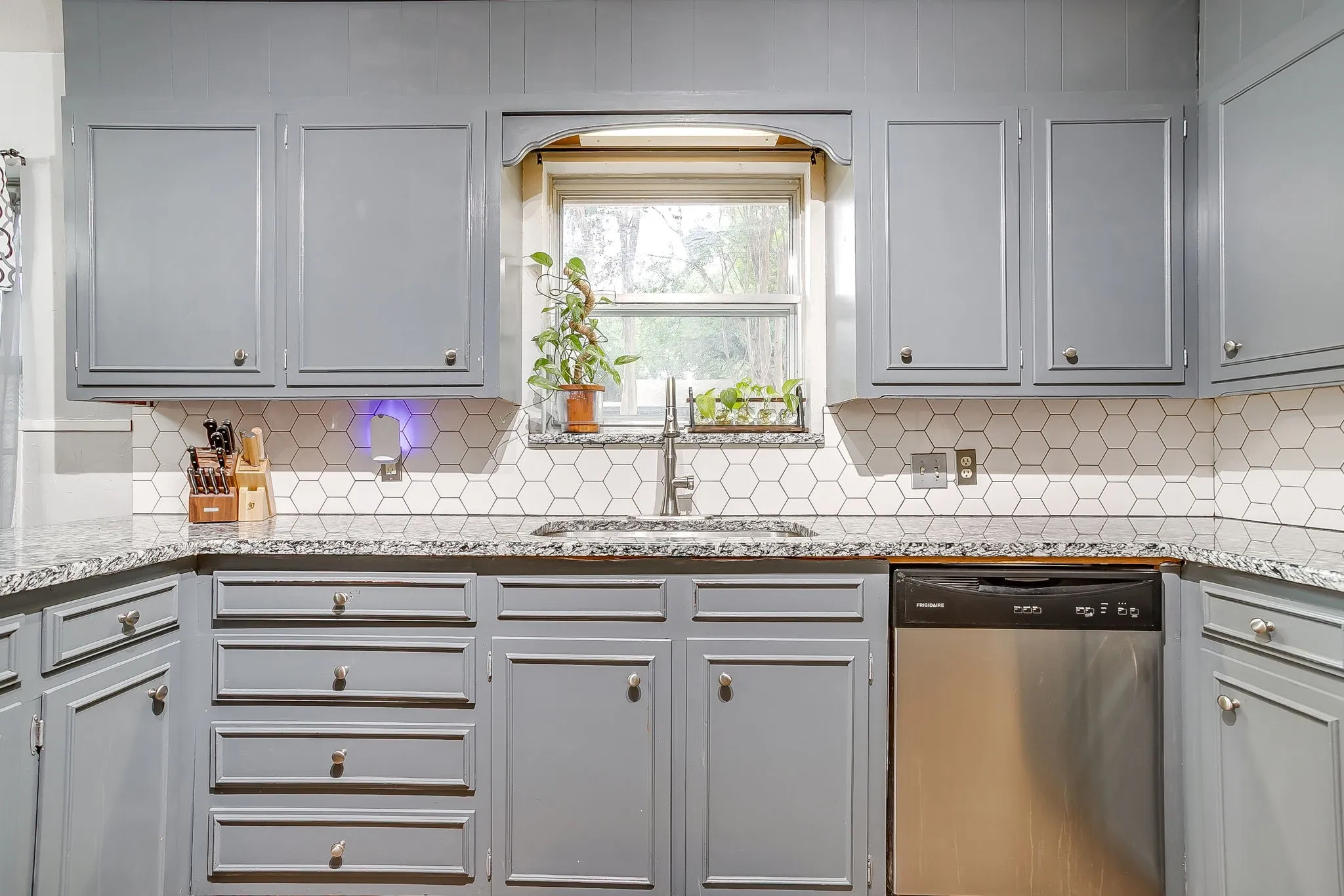 Kitchen featuring gray painted cabinetry, white hexagonal tile backsplash, granite countertops, and a stainless steel dishwasher