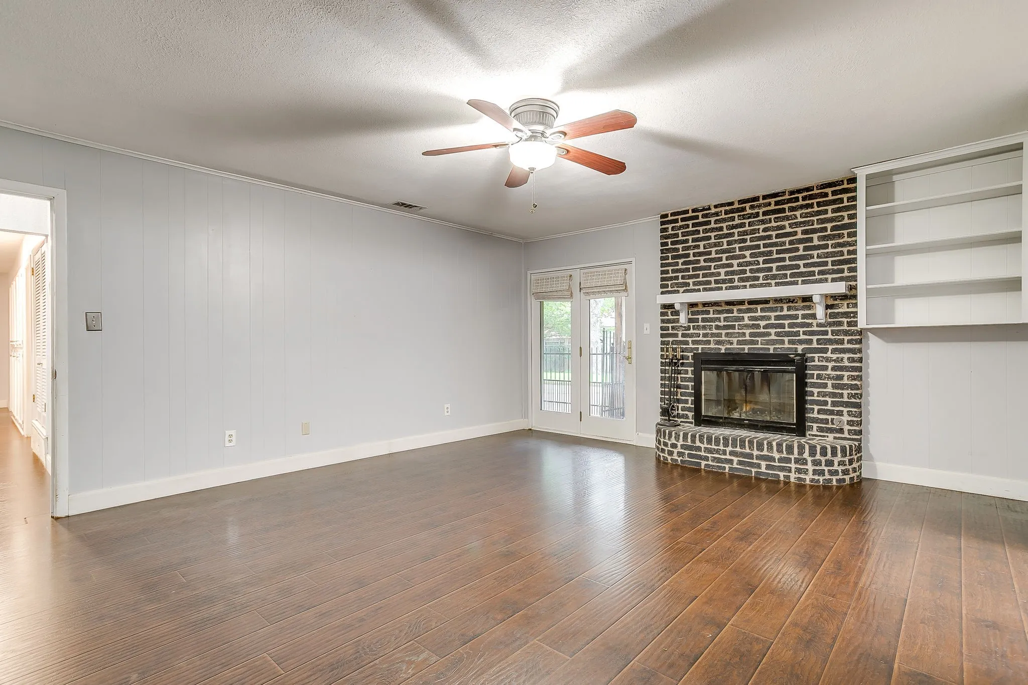 Spacious living area featuring wood-finish flooring, a ceiling fan with integrated lighting, and a brick fireplace with a white mantel and built-in shelving