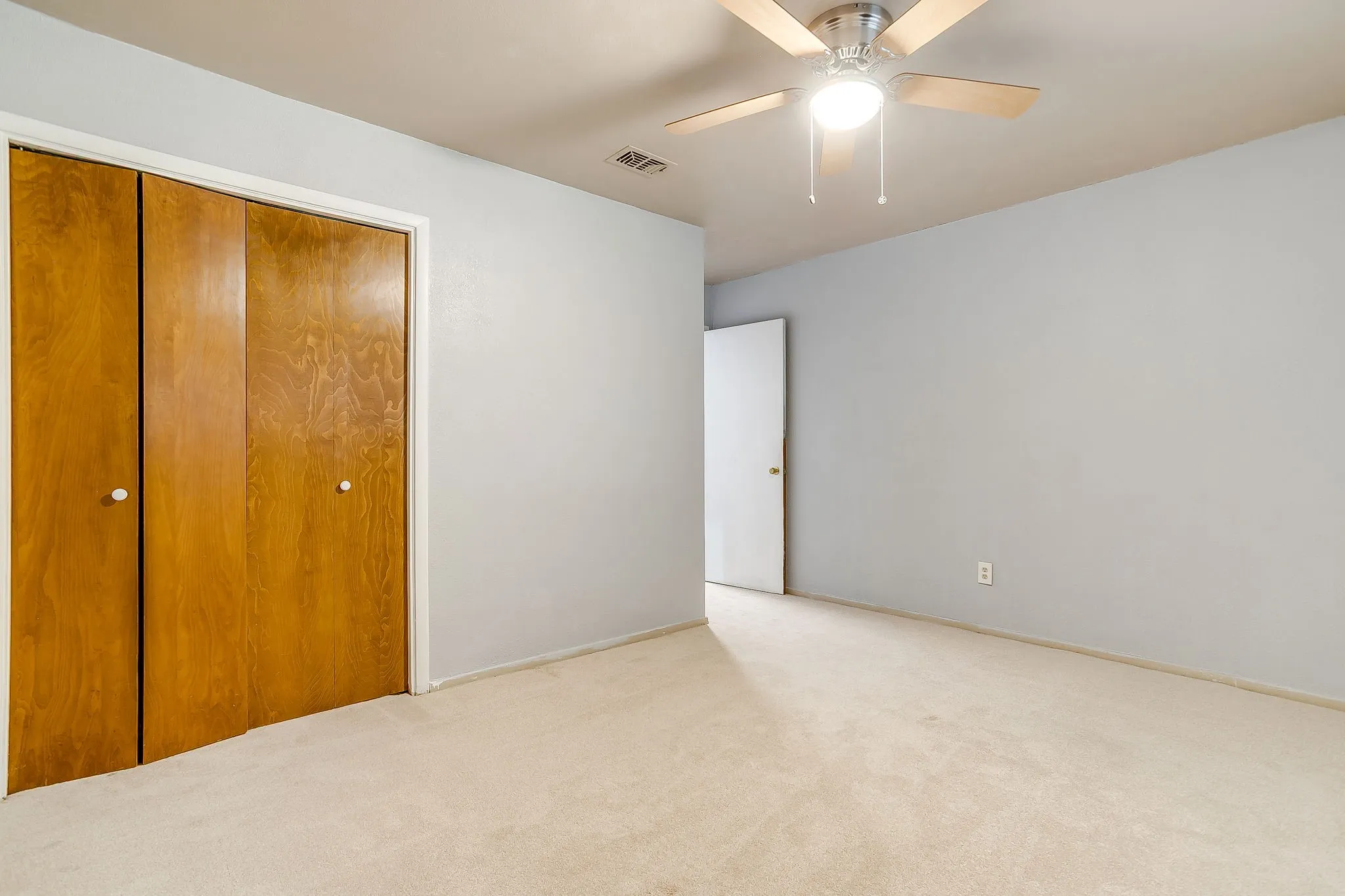 Neutral-toned carpeted room featuring a ceiling fan with light fixture, light gray walls, and wood-paneled bi-fold closet doors