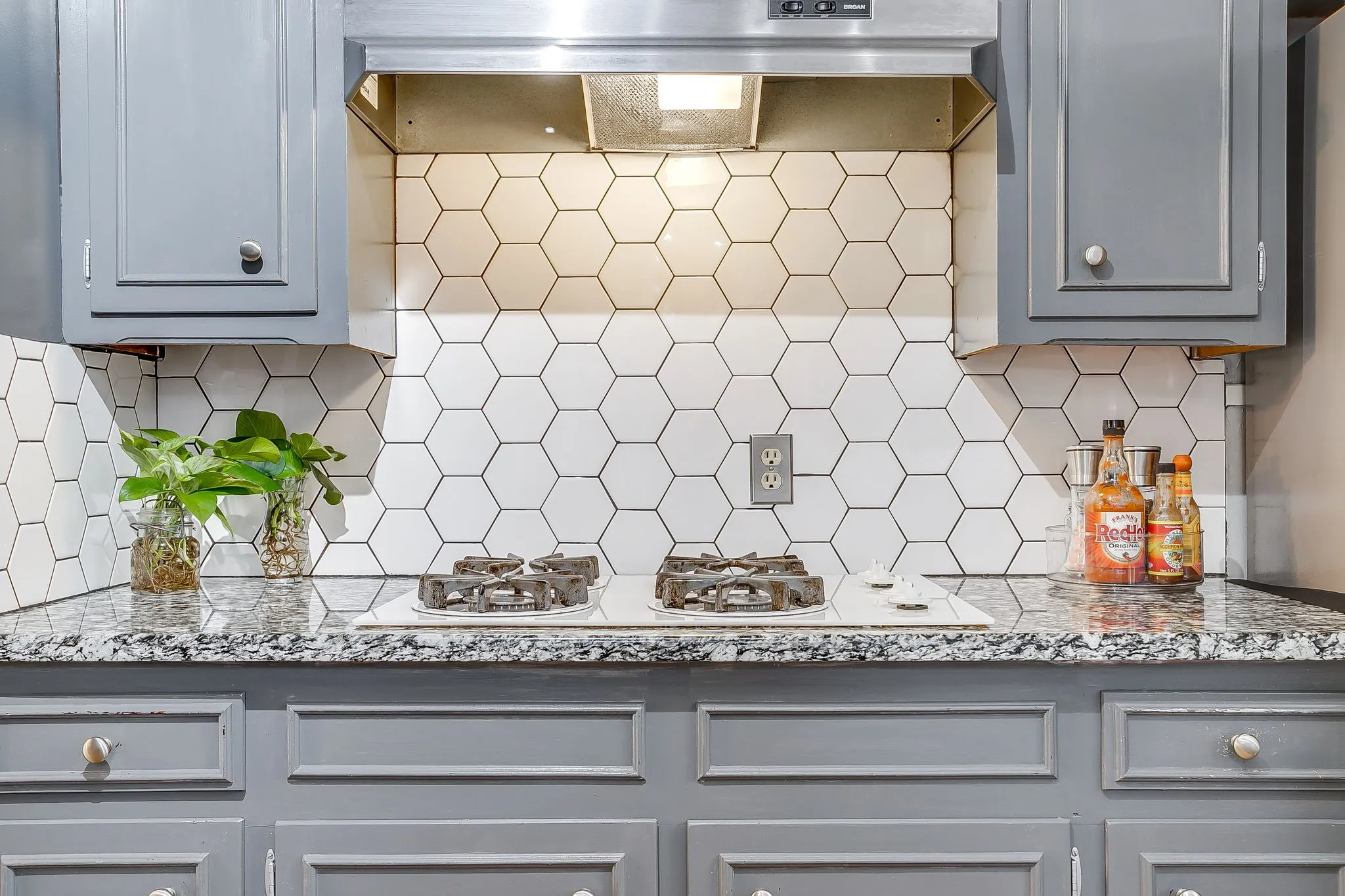 Kitchen featuring a built-in cooktop with an overhead stainless steel range hood, white hexagonal tile backsplash, light gray cabinetry with brushed nickel hardware, and speckled countertops