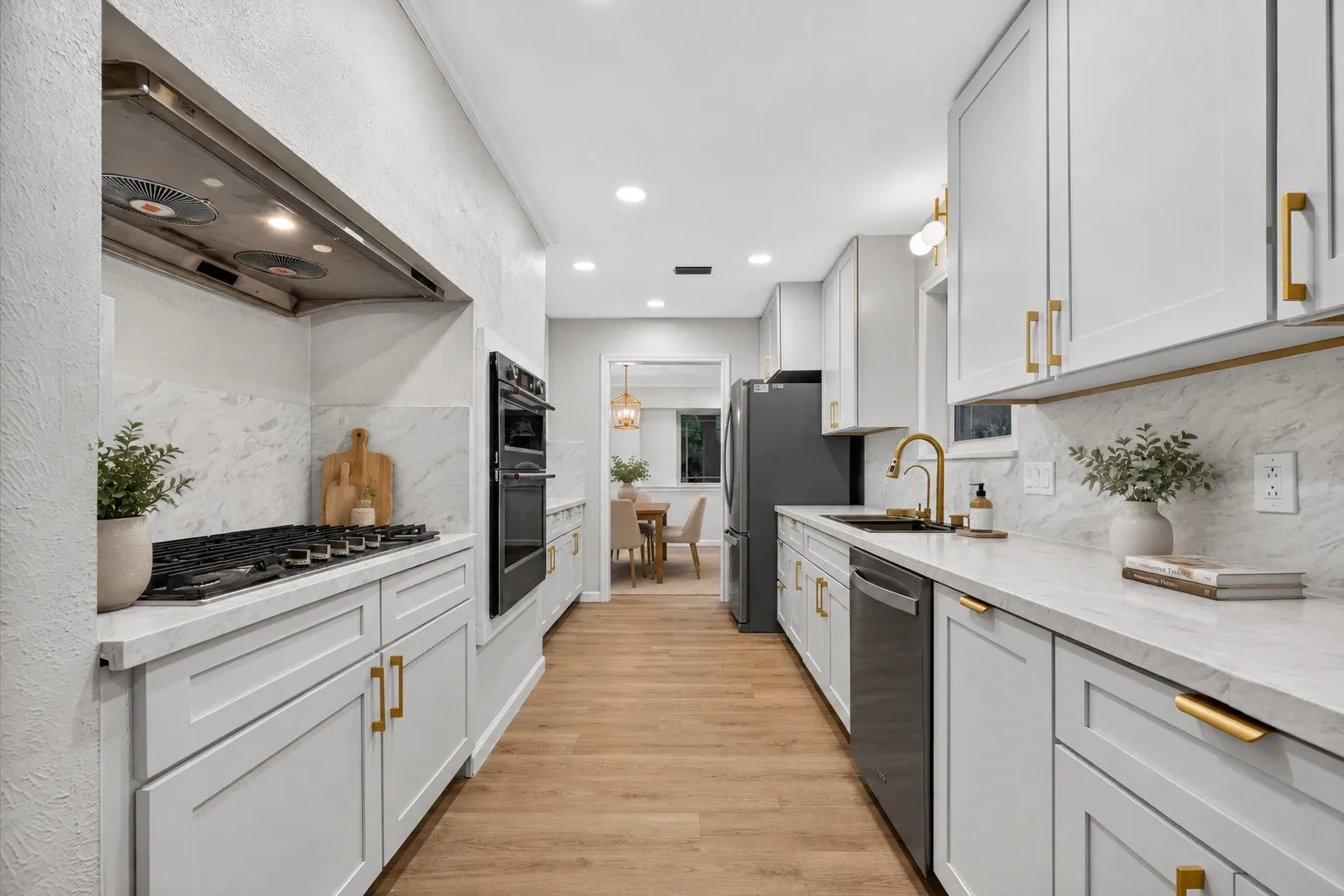 AI Staging: Galley kitchen featuring light wood-finish flooring, white cabinetry with gold-tone hardware, a gas range with a stainless steel vent hood, double wall ovens, and a gold-tone faucet