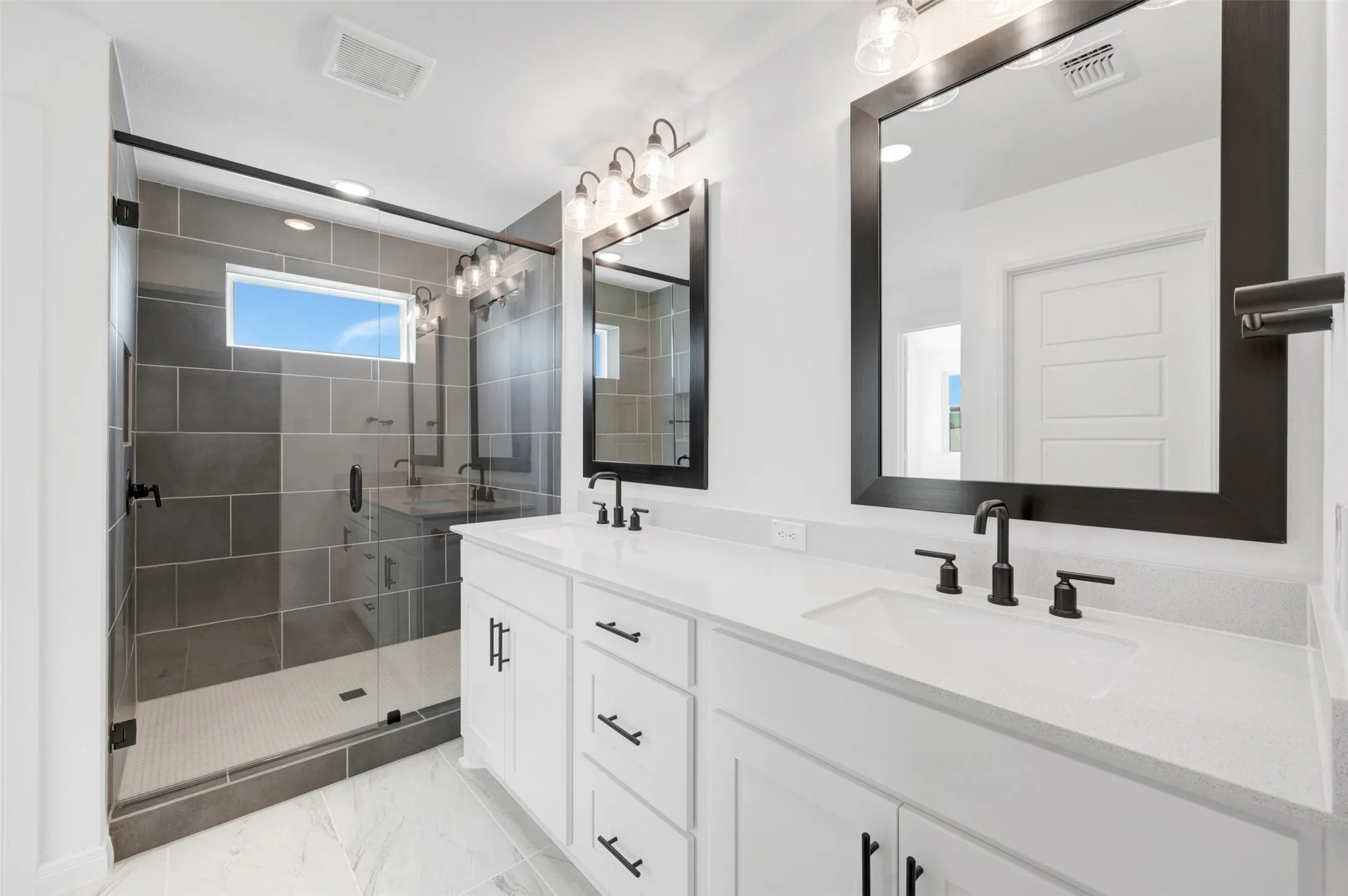 Dual vanity with white cabinetry and integrated sinks, matte black fixtures, framed mirrors, and a glass-enclosed shower with gray tile surround