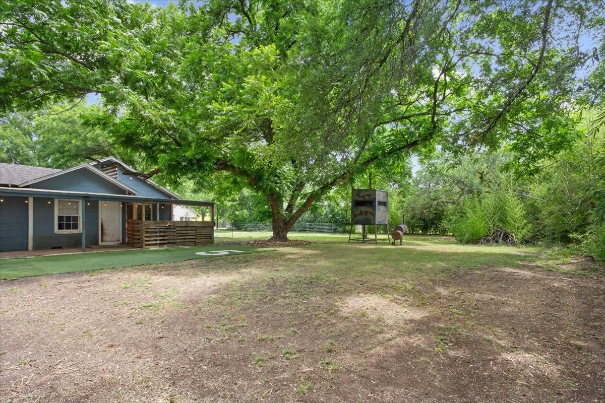 Spacious outdoor area featuring mature trees, a partially covered patio with string lights, and a gravel and grass ground cover