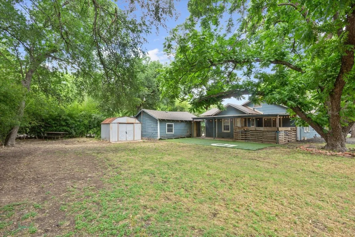 Expansive backyard featuring mature trees, a detached storage shed, a separate outbuilding, a covered porch with slatted wood railing, and a section of artificial turf