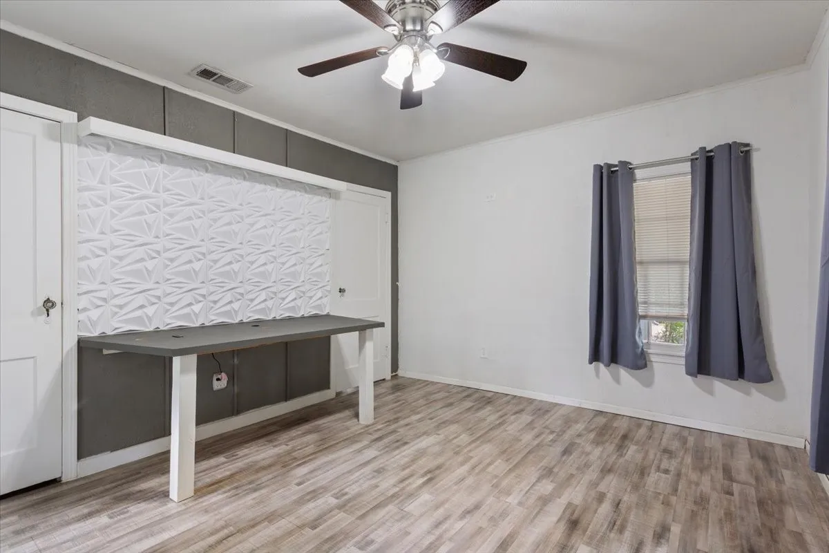 Room featuring wood-finish flooring, a built-in desk with a textured accent wall, two white paneled doors, and a ceiling fan with integrated lighting