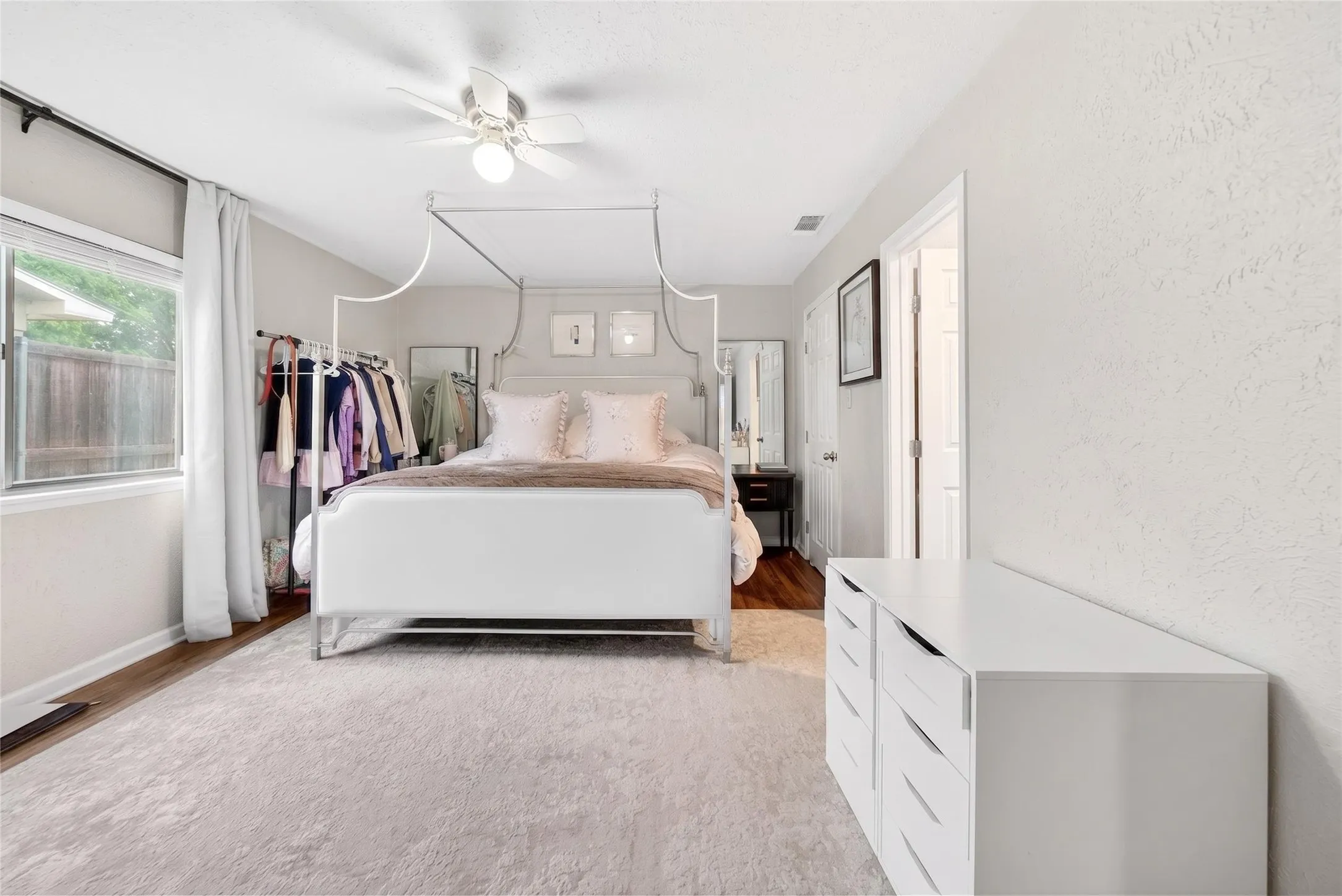 Spacious bedroom featuring wood-finish flooring, a ceiling fan, and a large window with white curtains