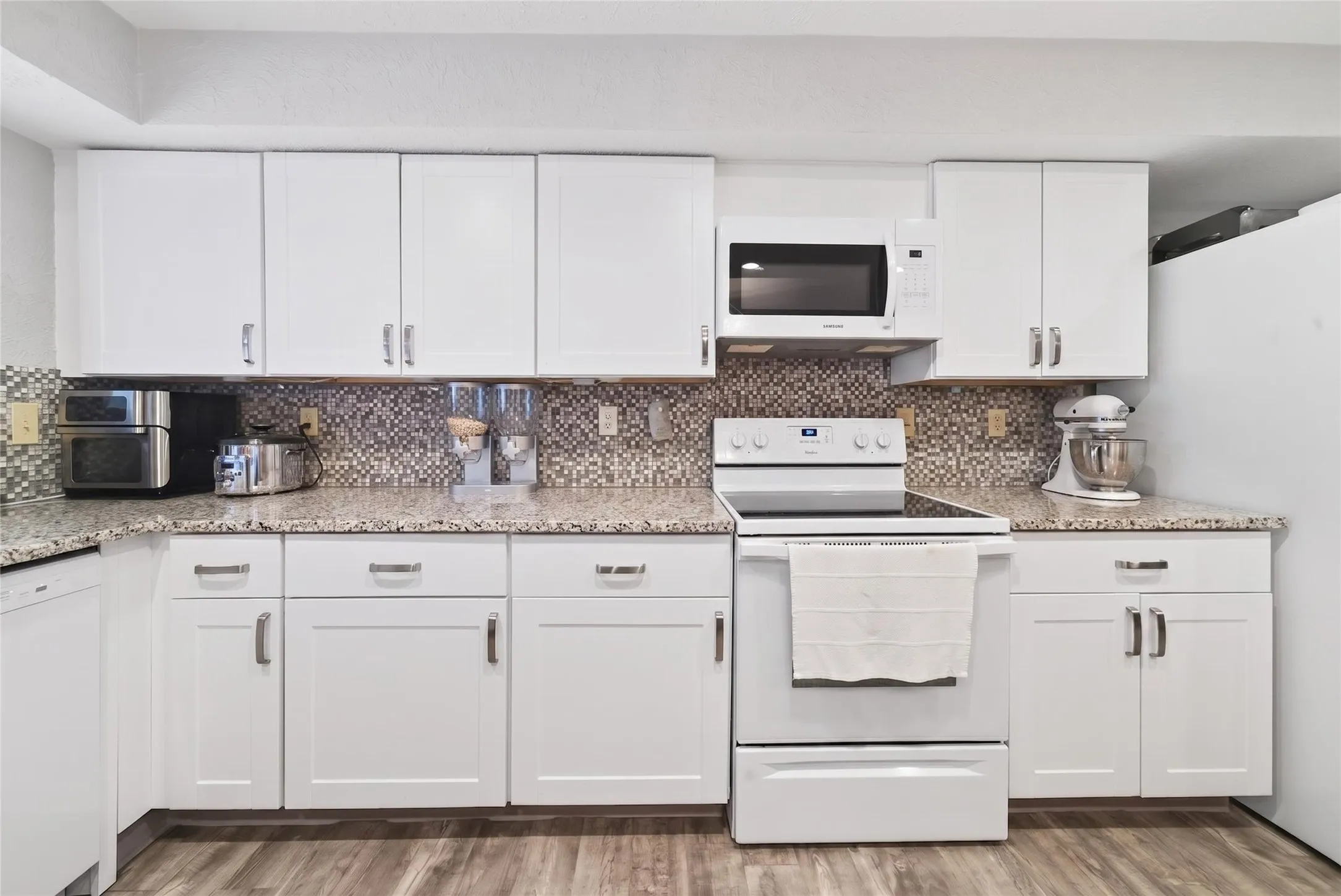 Modern kitchen featuring white cabinetry, a mosaic tile backsplash, speckled countertops, a built-in microwave, and wood-finish flooring