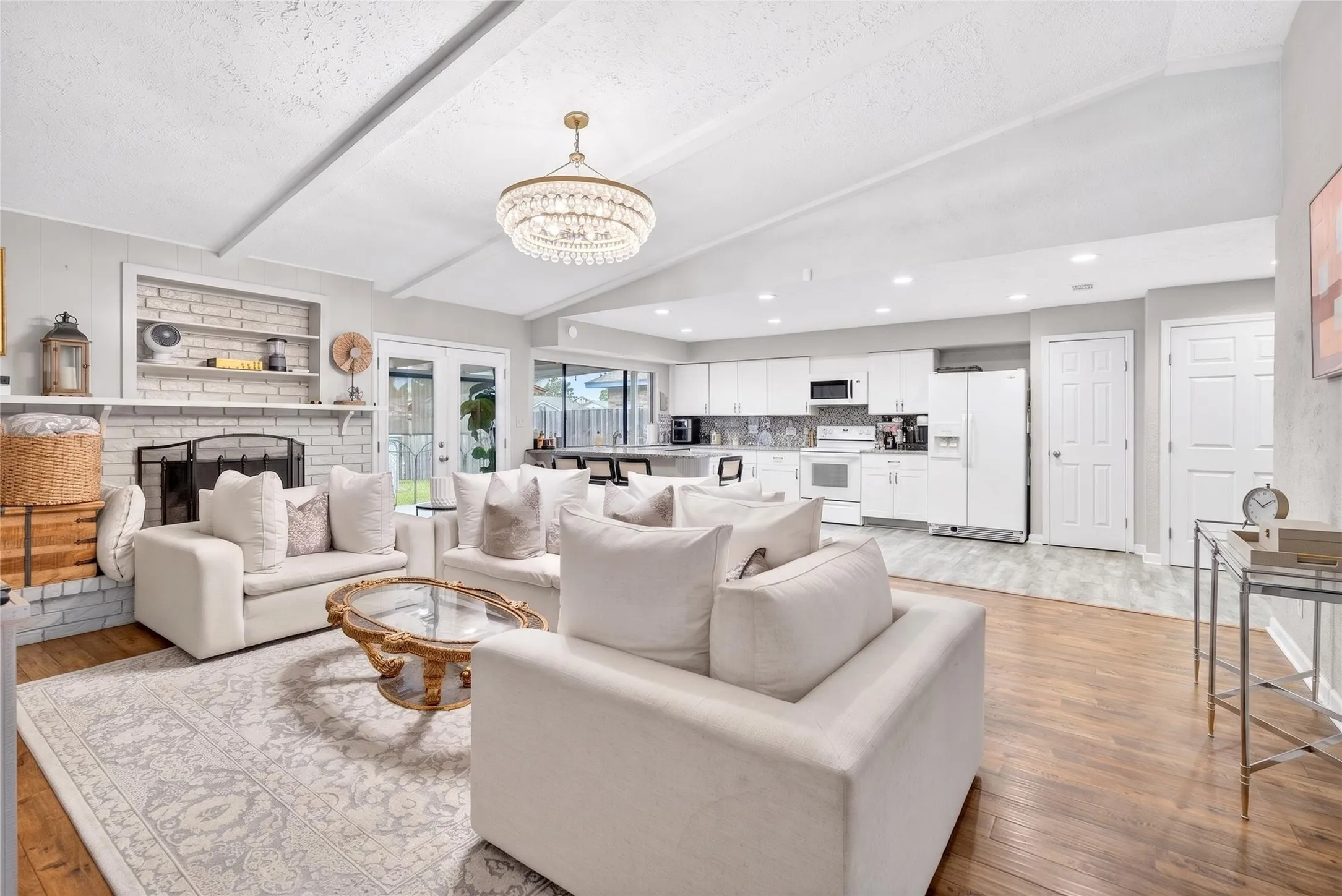 Spacious living area featuring vaulted ceilings with exposed beams, a white brick fireplace with built-in shelving, wood-finish flooring, and an open-concept kitchen with white cabinetry