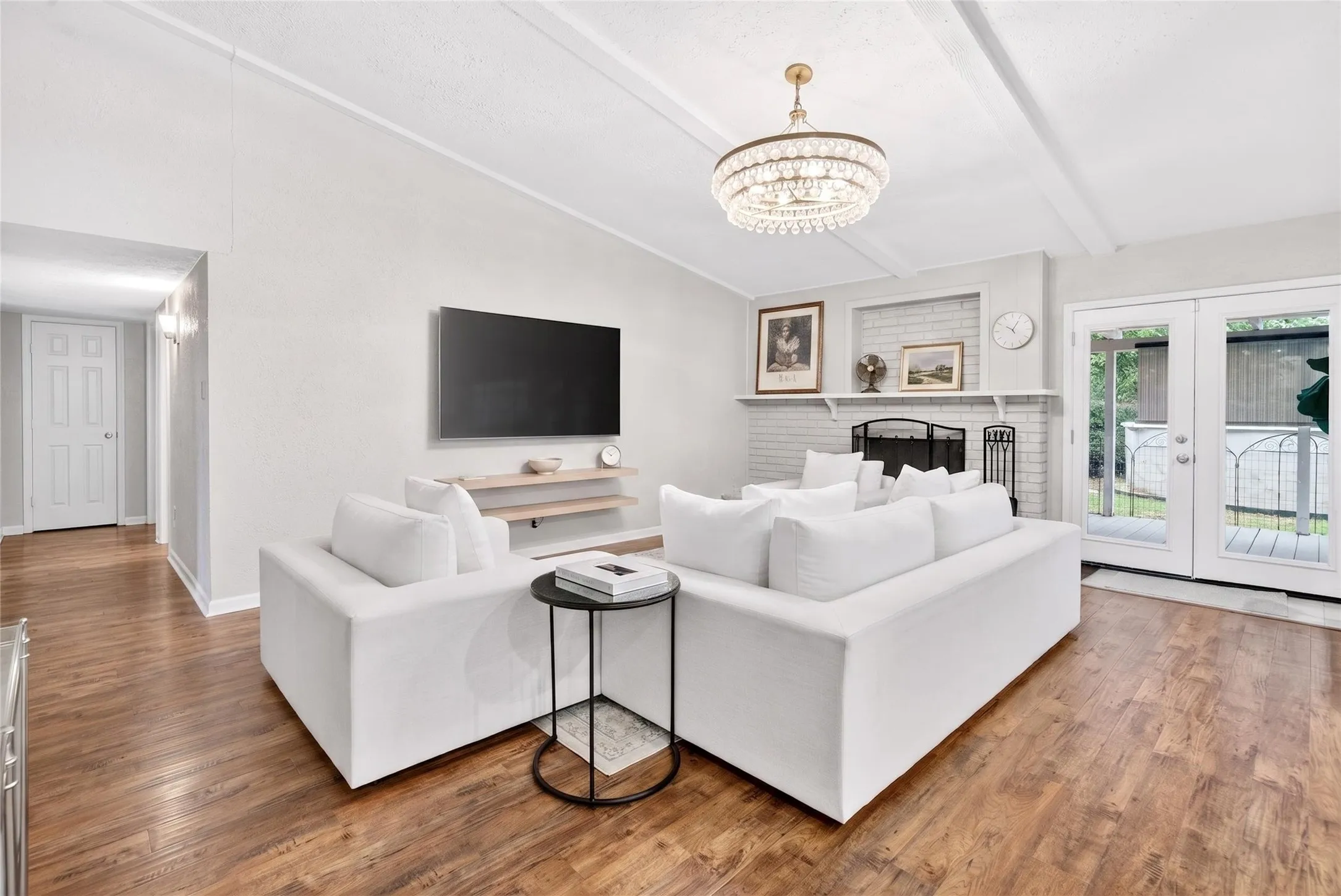 Spacious living area featuring wood-finish flooring, a white brick fireplace with mantel, exposed ceiling beams, and French doors providing exterior access