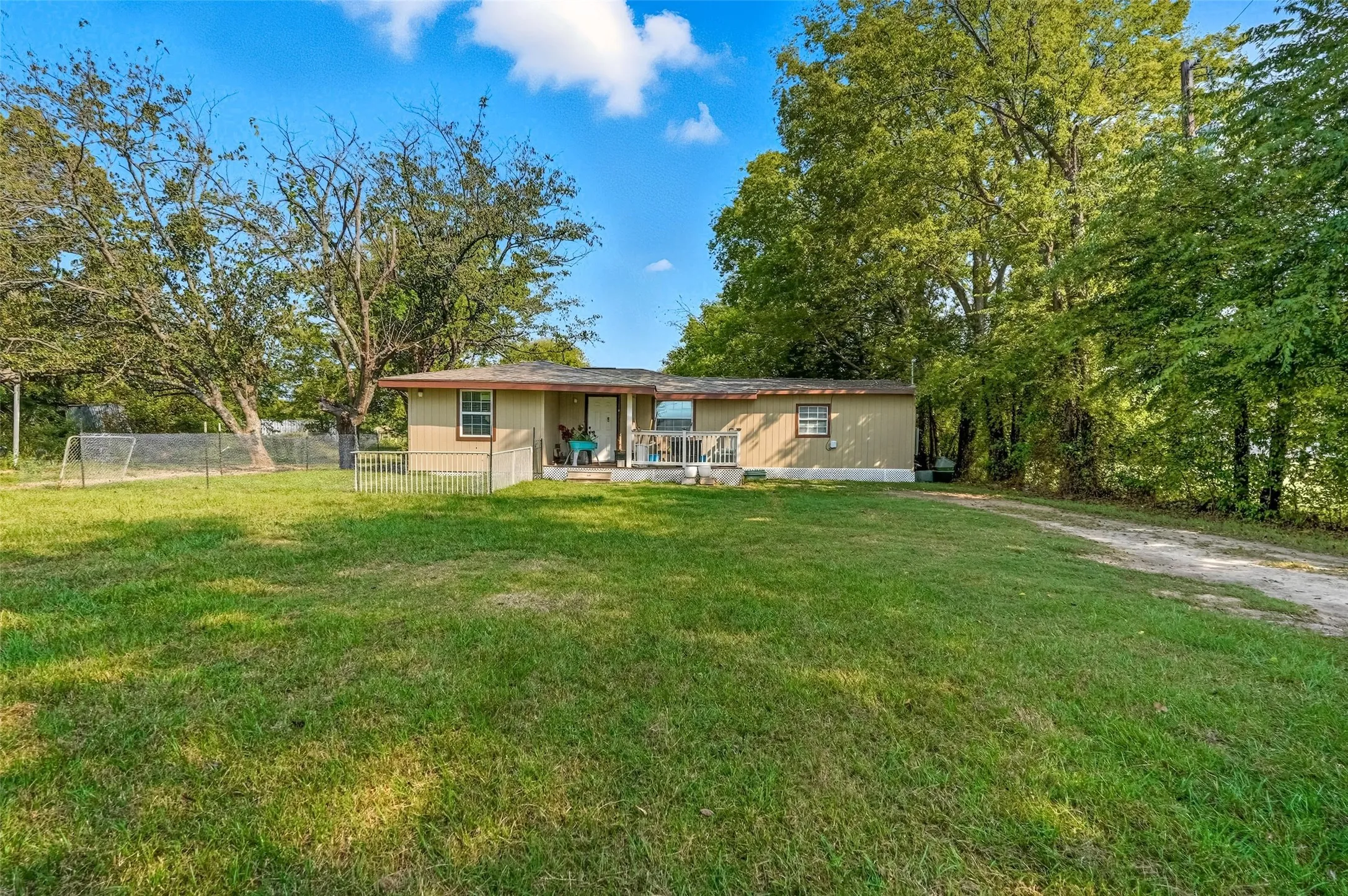 Expansive front yard with a large grassy area, mature trees, and a dirt driveway leading to the residence