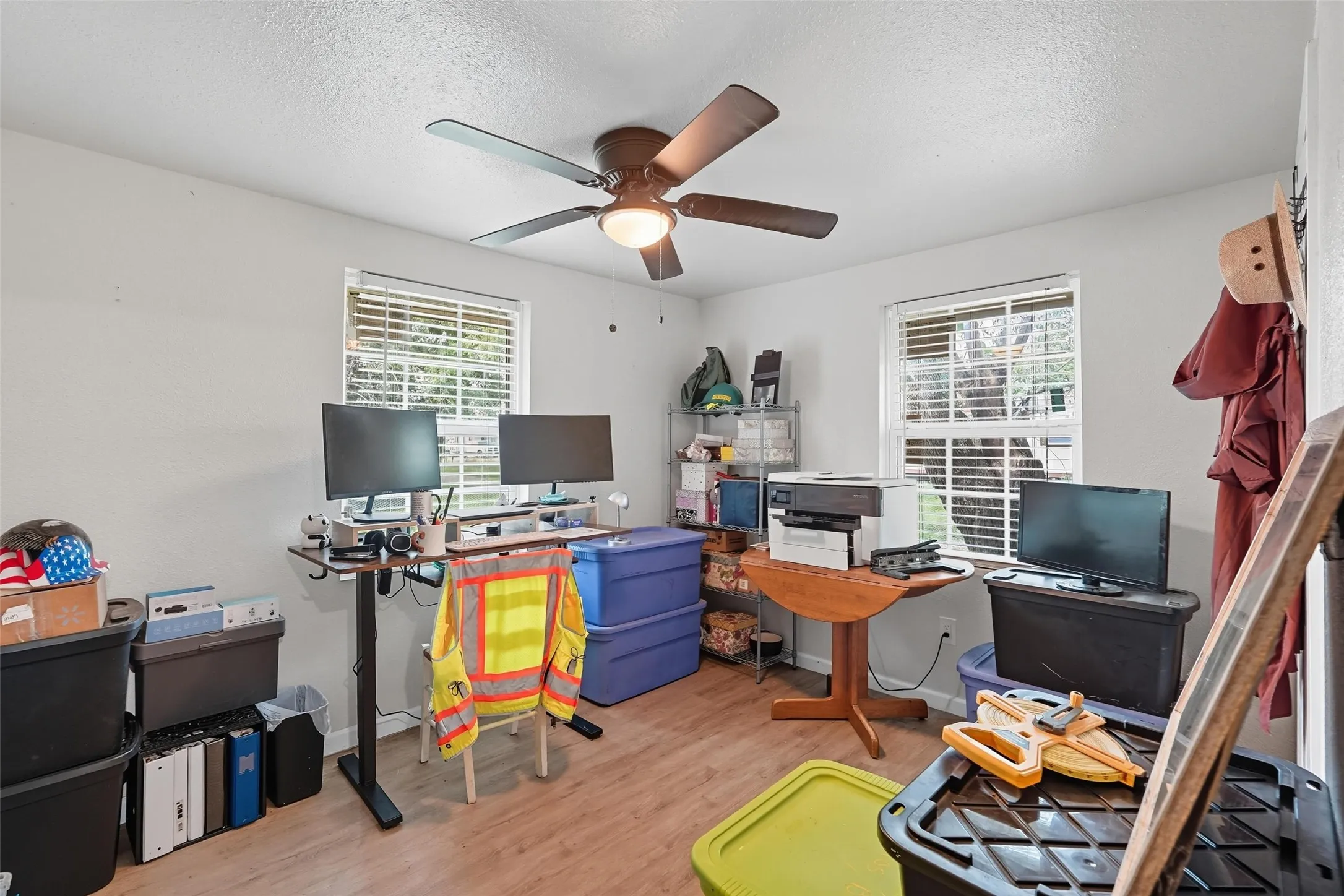 Room with wood-finish flooring, two large windows with blinds, a ceiling fan with integrated lighting, and light-colored walls