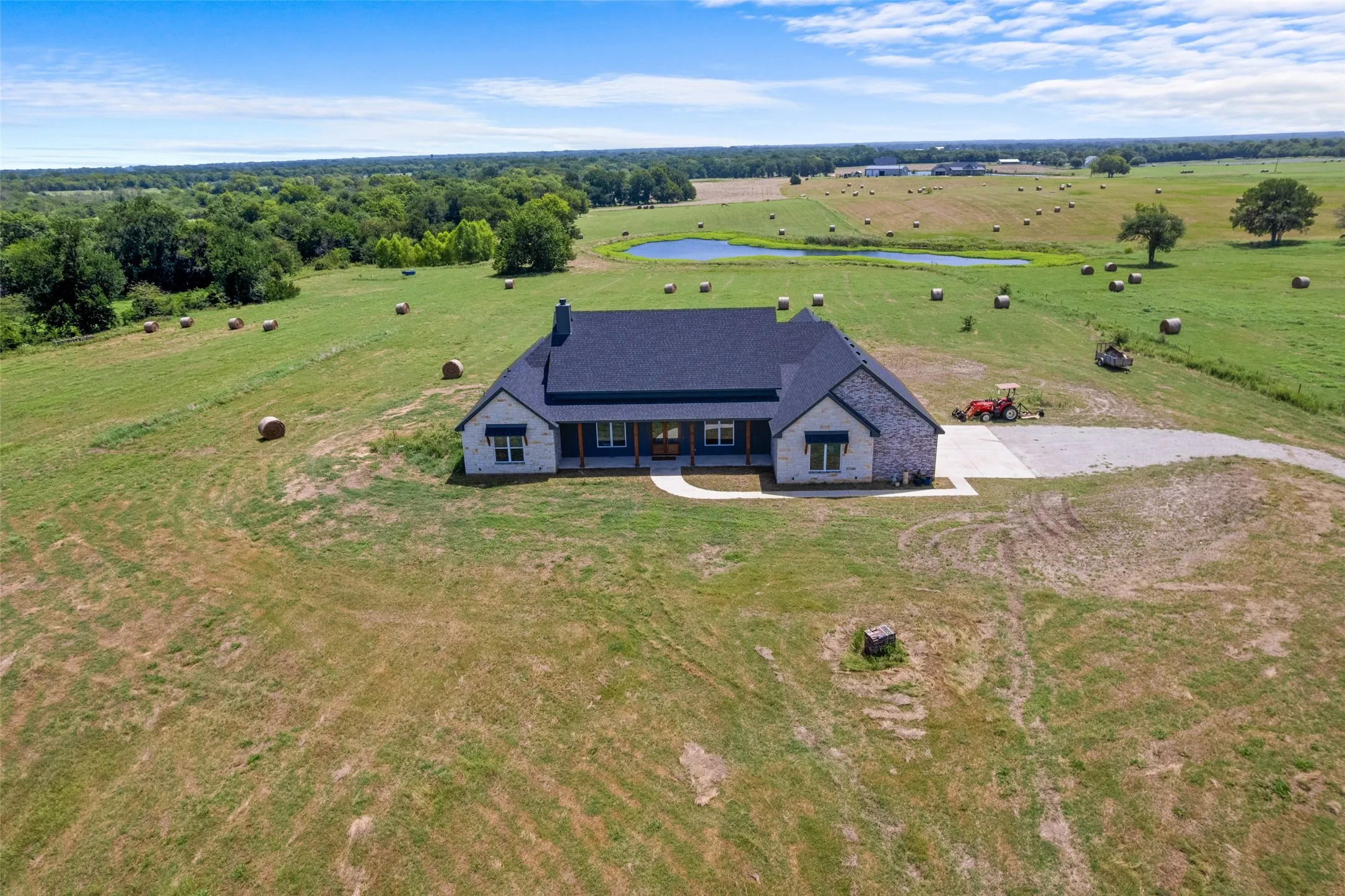 Expansive rural property featuring a stone and siding residence with a dark roof, surrounded by open fields, trees, and water features
