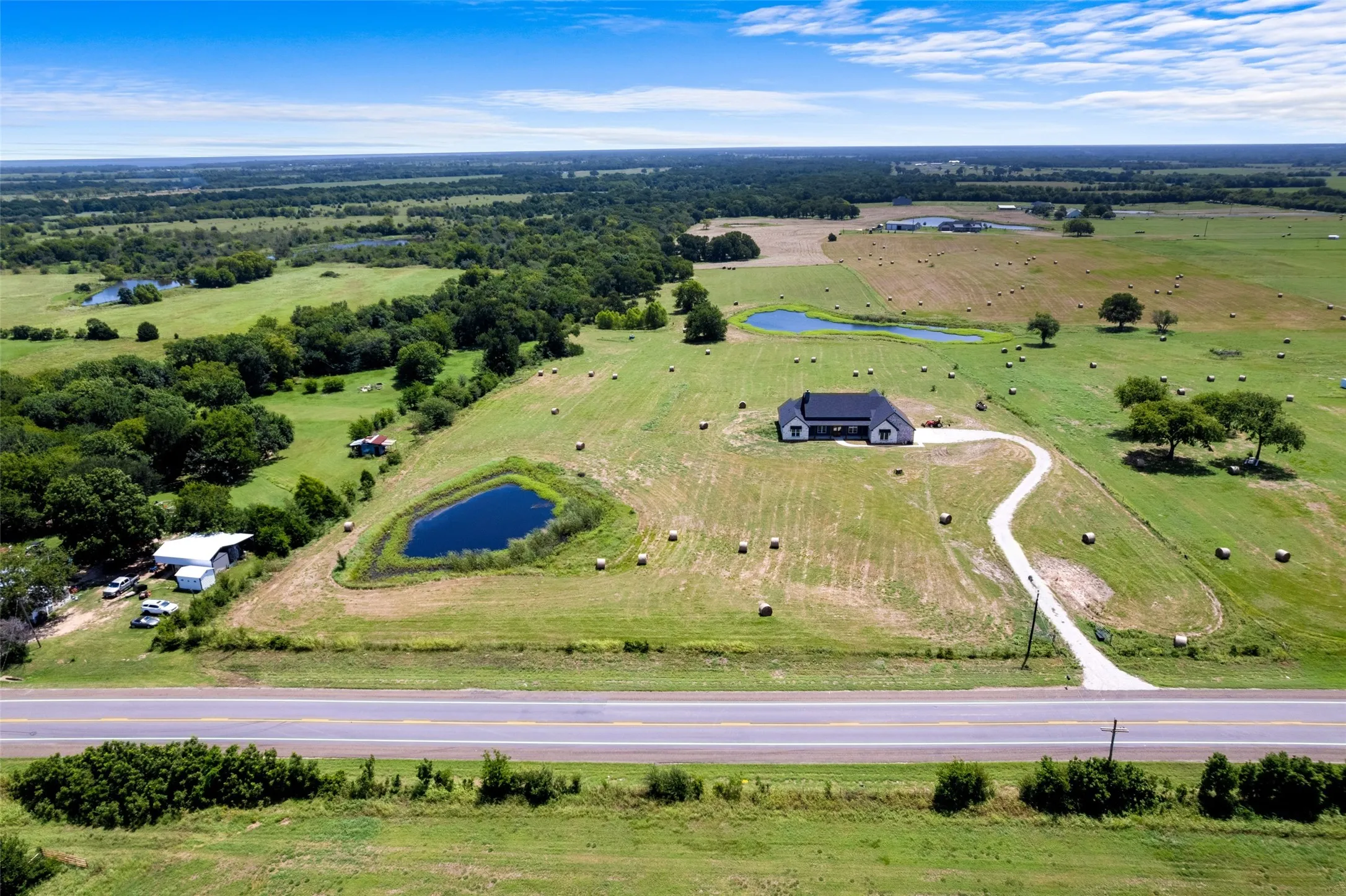 Expansive rural property featuring a modern single-story residence with a dark roof, a private pond, mature trees, and a winding driveway