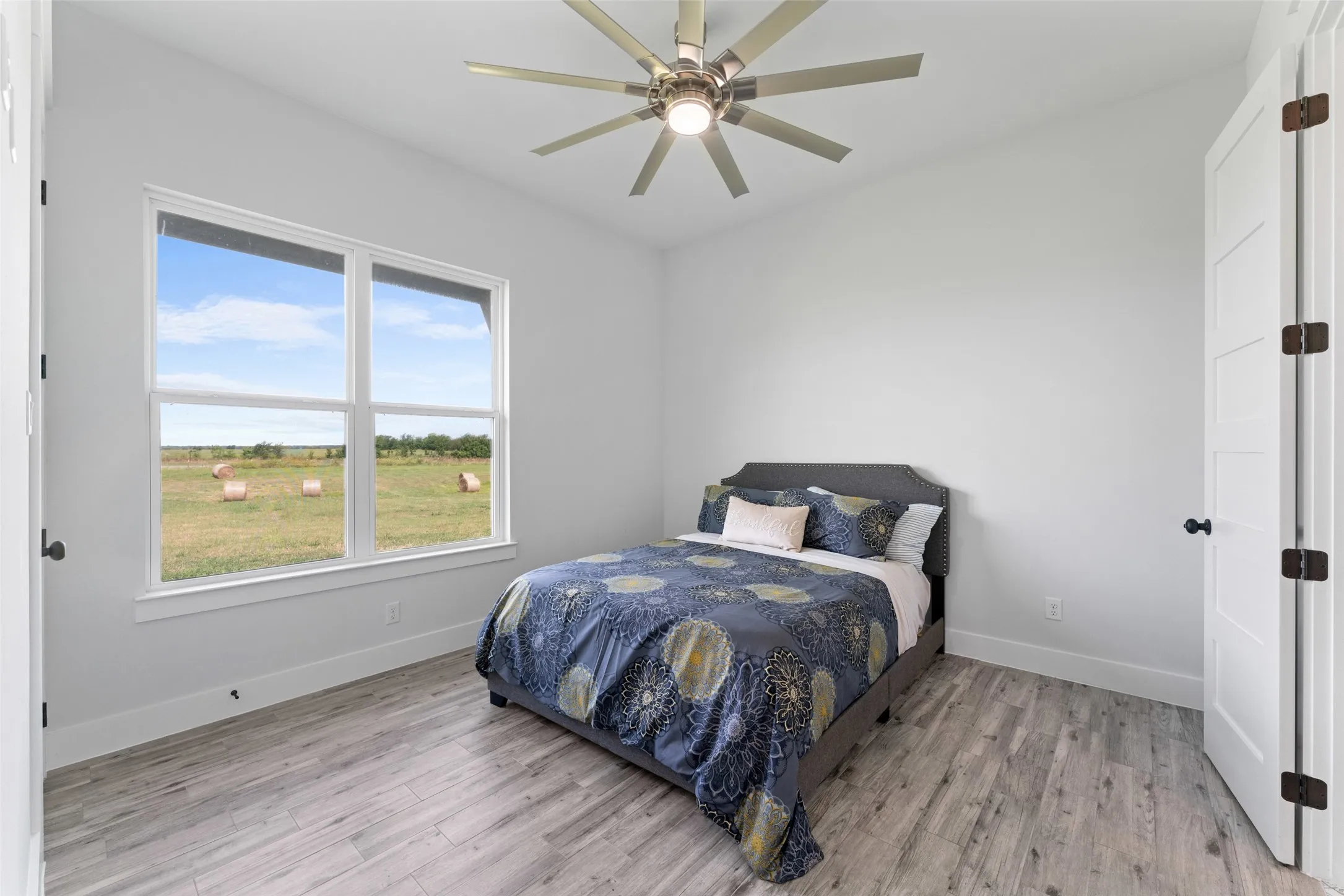 Bright interior space featuring a large window with white trim, wood-finish flooring, a contemporary ceiling fan with integrated lighting, and a white paneled door with dark hardware