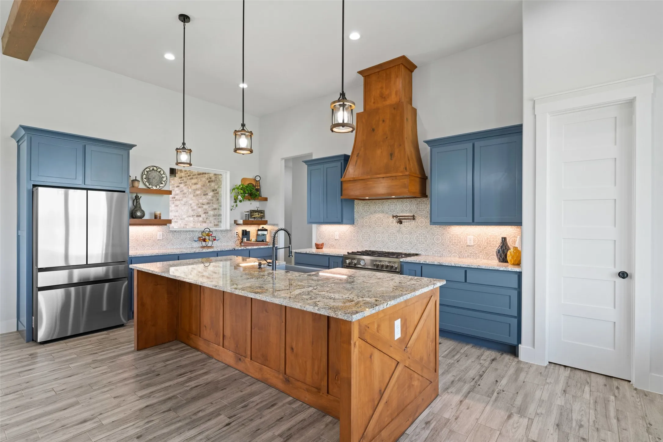 Spacious kitchen featuring a large wood-finish island with a stone-finish countertop, blue cabinetry, a wood-finish range hood, stainless steel appliances, and light wood-finish flooring
