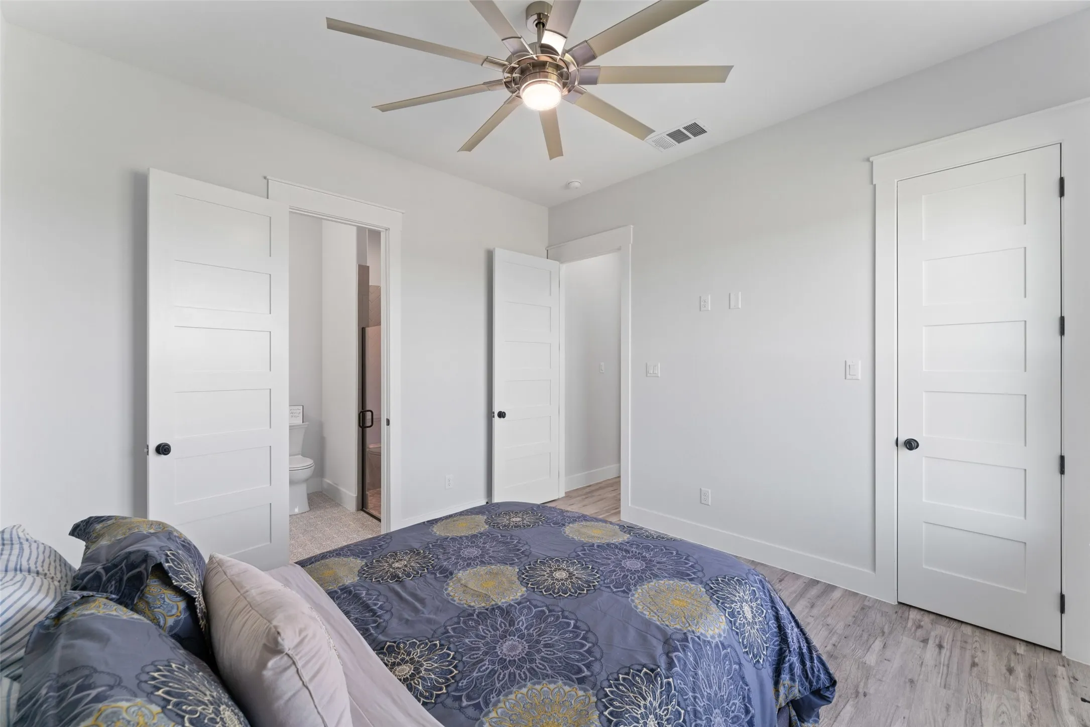 Bedroom featuring an overhead ceiling fan with lighting, wood-finish flooring, white paneled doors with black hardware, and an adjacent bathroom with a toilet and shower enclosure