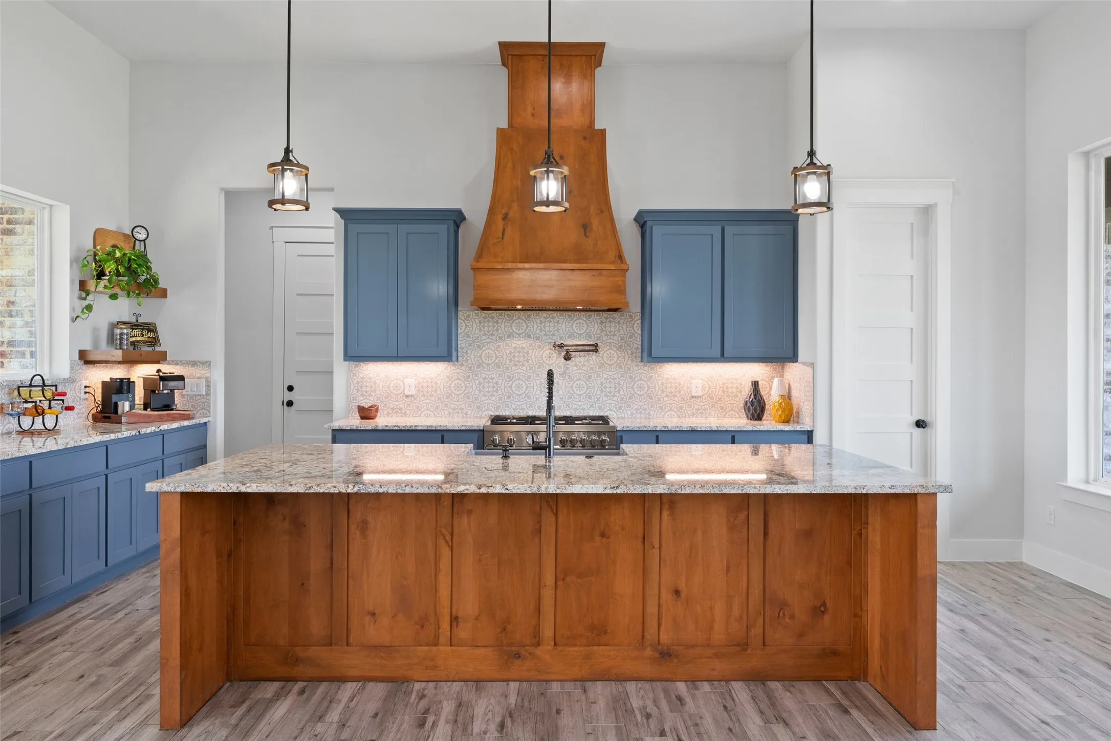 Spacious kitchen featuring a large wood-finish island, gray-flecked countertops, blue cabinetry, a decorative wood-finish range hood, and pendant lighting