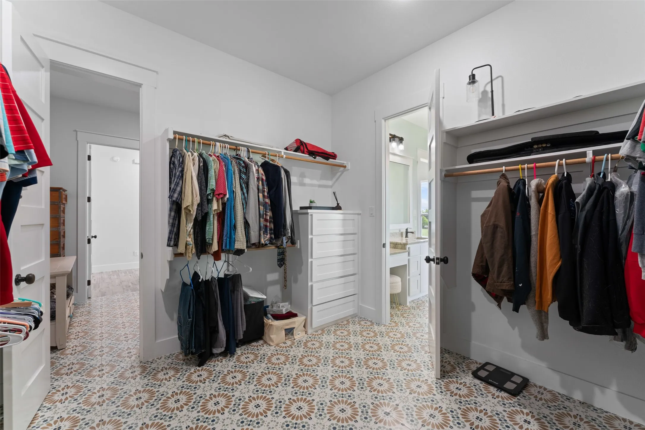 Spacious walk-in closet featuring patterned floor tiles, extensive built-in shelving, hanging rods, a white dresser, and a doorway opening to a bathroom with a vanity