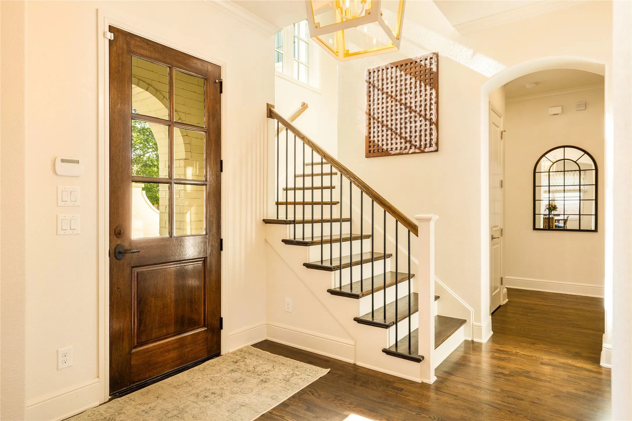 Bright and welcoming entryway showcases rich wood flooring, elegant arched details, and a striking staircase with classic iron balusters. Natural light pours through oversized windows and the glass paneled front door, creating a warm and sophisticated first impression.