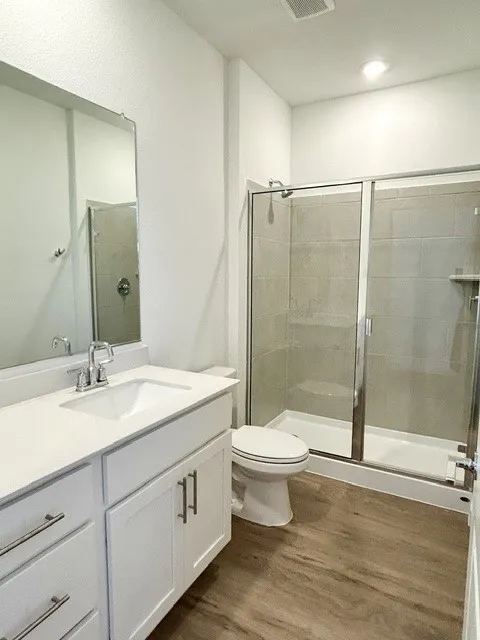 Bathroom featuring a single vanity with white cabinetry and a light-colored countertop, chrome fixtures, a mirrored wall, a glass-enclosed shower, and wood-finish flooring