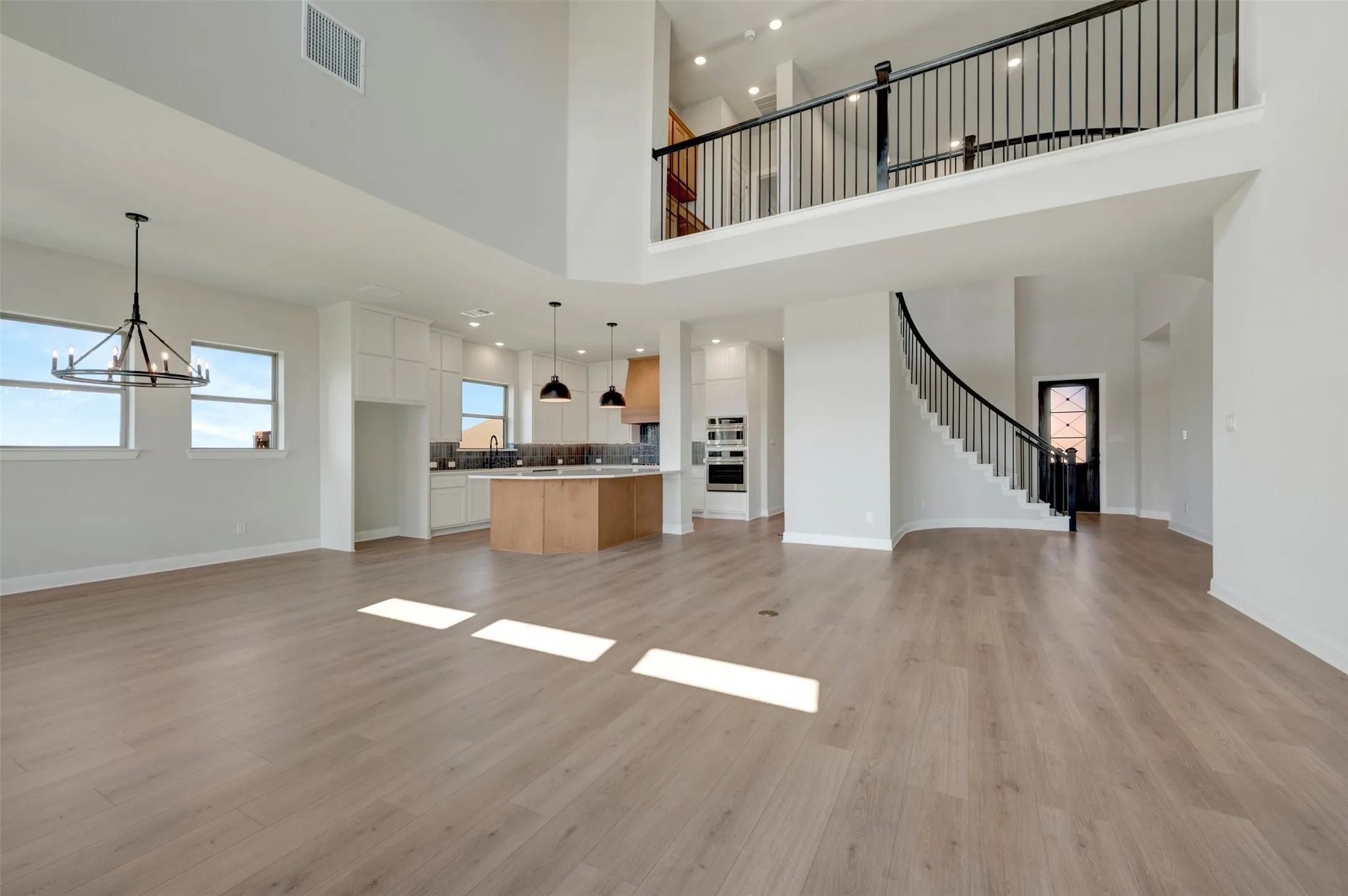 Open-concept living area with wood-finish flooring, a contemporary kitchen featuring an island, high ceilings, a curved staircase with black railings, and a second-story landing
