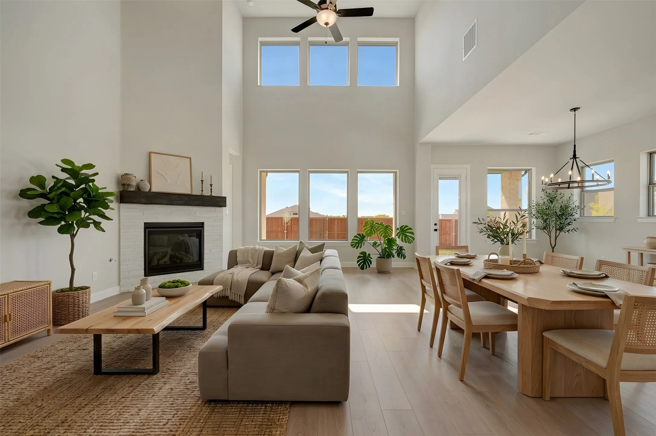Vaulted ceiling with a ceiling fan, white brick fireplace with a dark wood-finish mantel, light wood-finish flooring, multiple windows, and a black chandelier