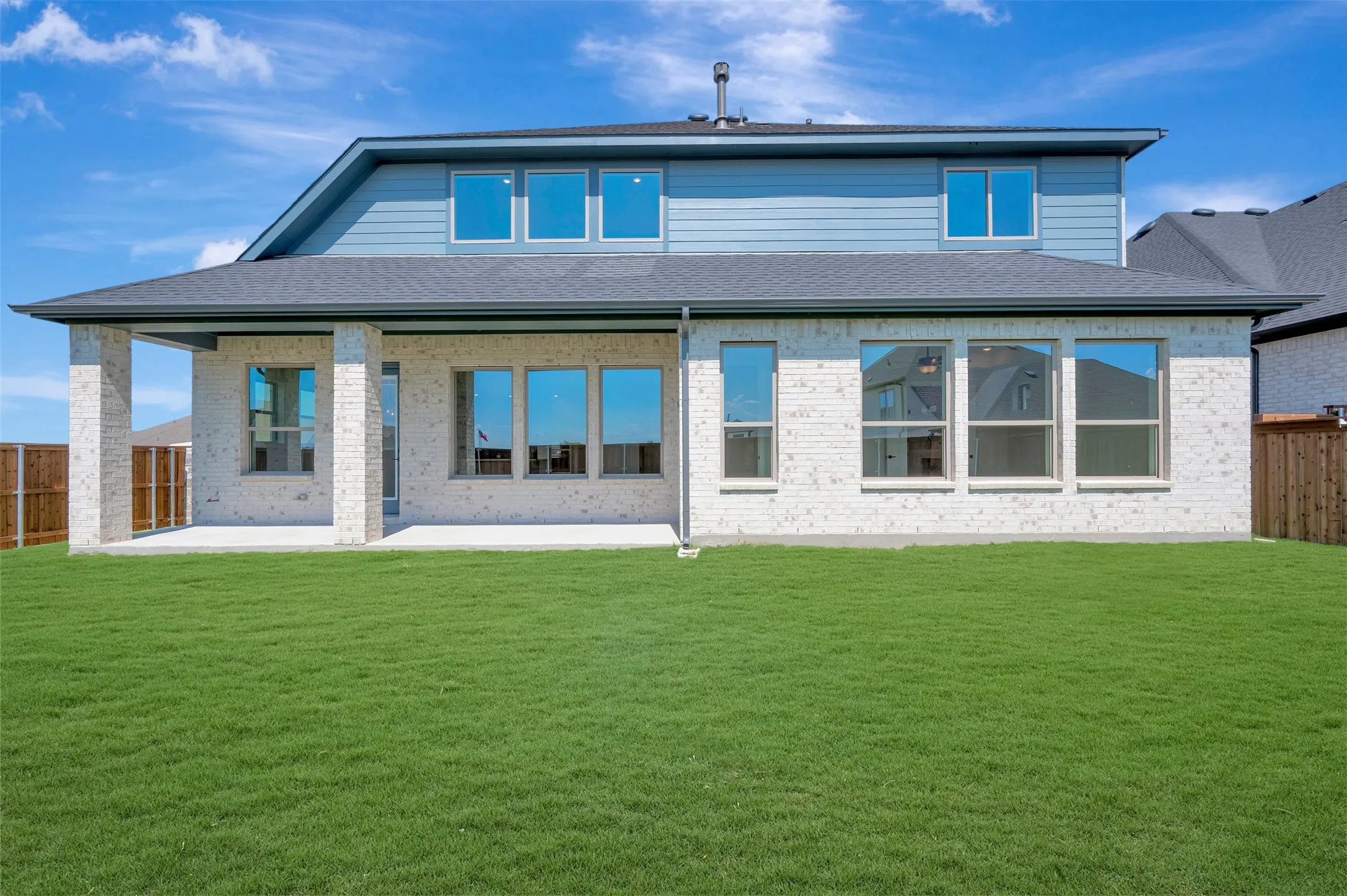 Expansive green lawn with a covered patio featuring stone-look columns, multiple large windows, and a two-story exterior with light-colored siding