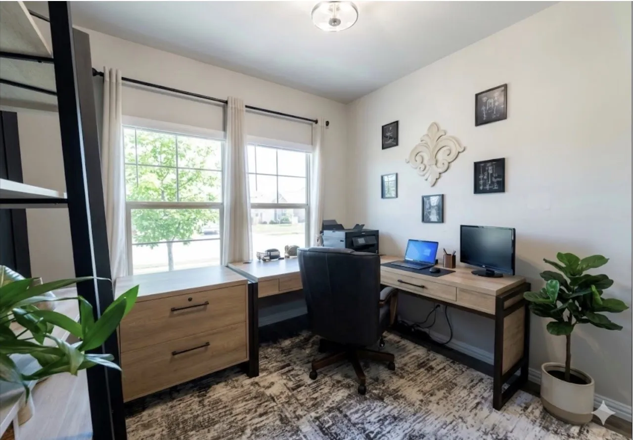 Office space featuring an L-shaped desk, a large window, a ceiling-mounted light fixture, light-toned wall paint, and patterned area rug