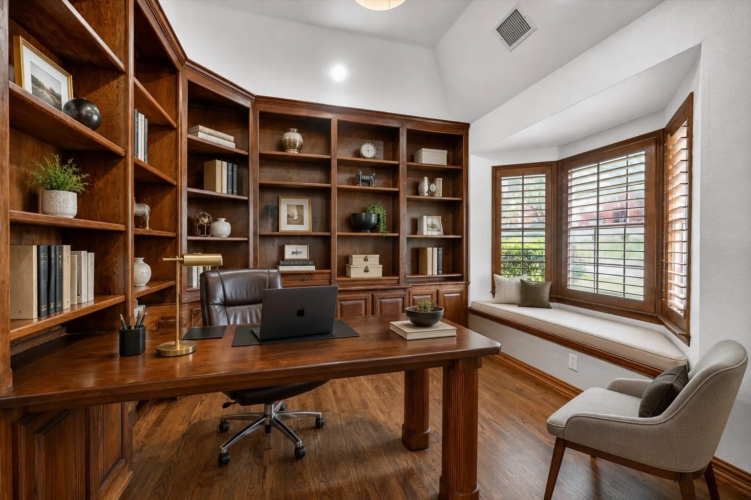 Dedicated office space featuring extensive built-in wood bookcases, wood-finish flooring, a bay window with integrated seating, plantation shutters, and a vaulted ceiling