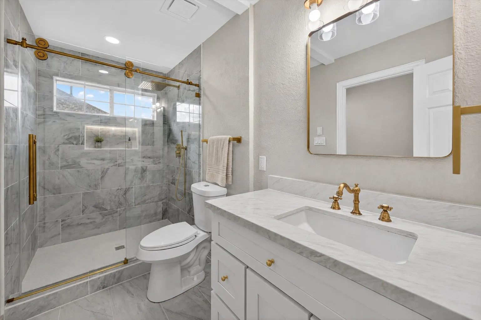 Bathroom featuring a glass-enclosed shower with gold-tone hardware, a tiled shower surround with a recessed niche, a white vanity with a rectangular undermount sink, a stone-finish countertop, and gold-tone vanity faucet