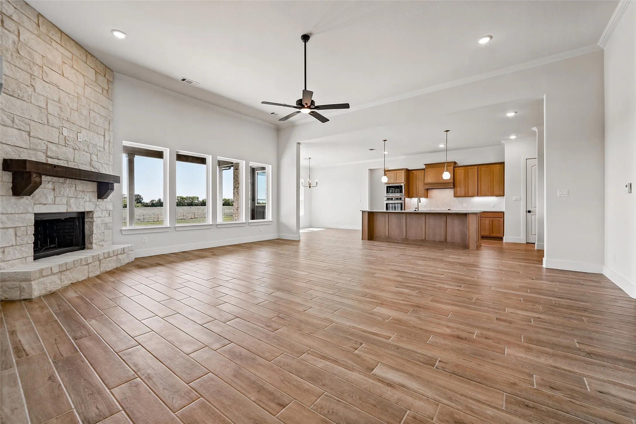 Spacious open-concept living area featuring a stone-clad fireplace with a wood mantel, wood-finish tile flooring, and multiple large windows