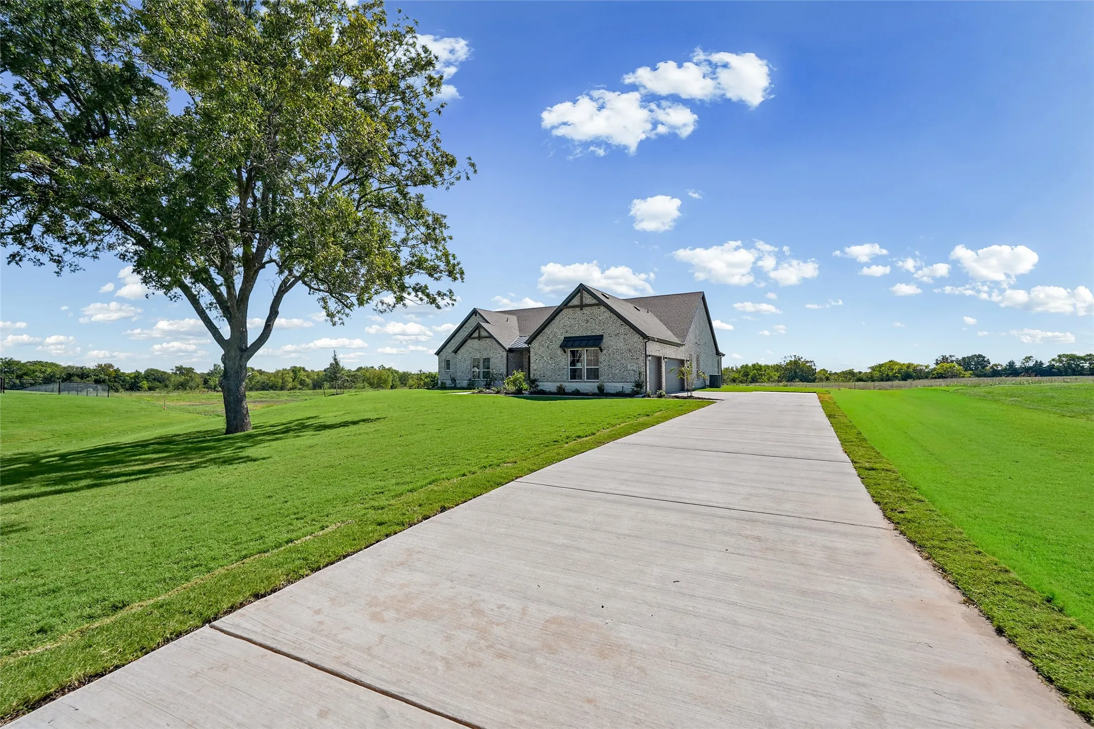 Contemporary stone exterior with a dark-pitched roof, large concrete driveway, expansive grassy lawn, and mature tree