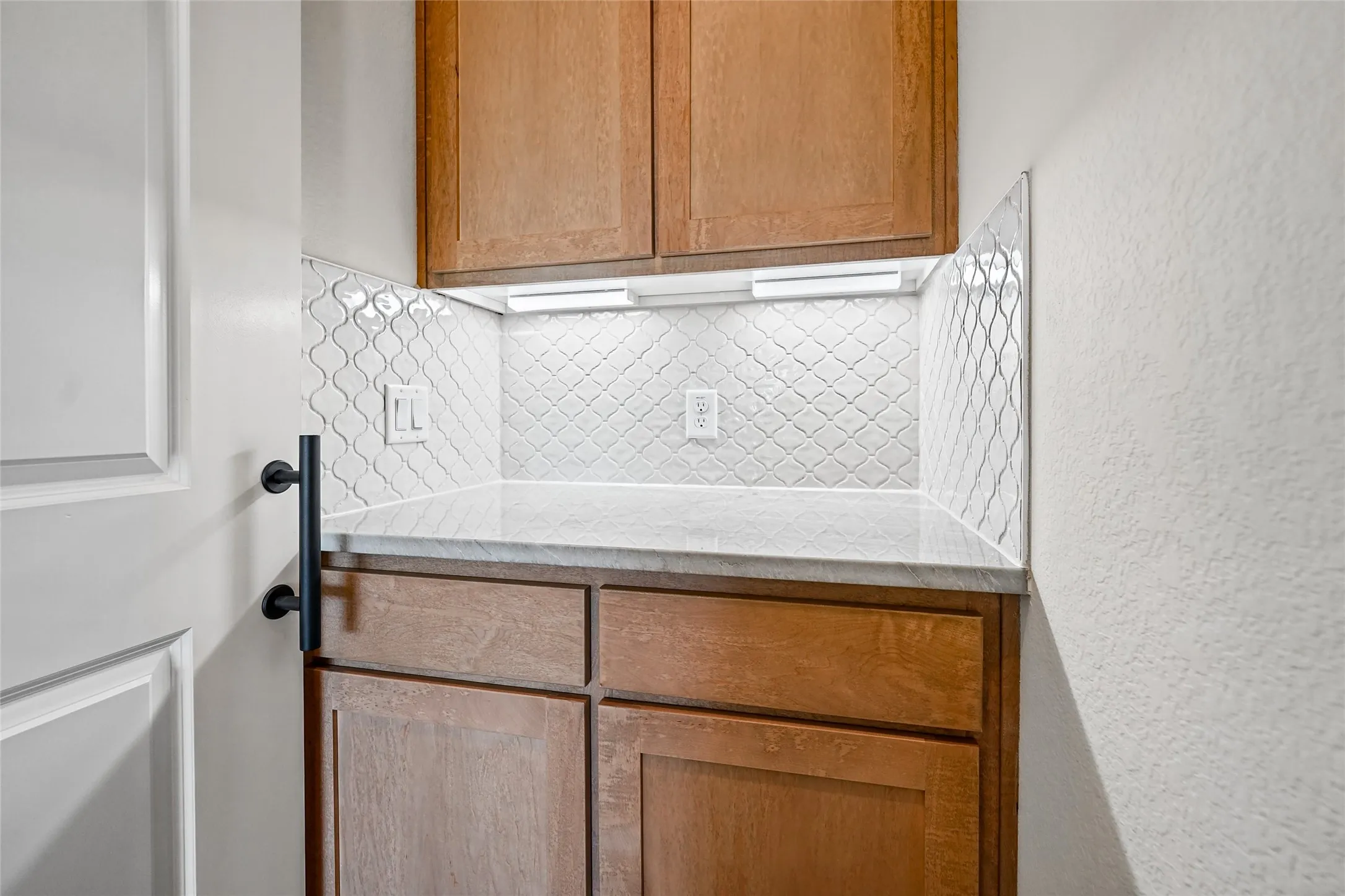 Built-in cabinetry with wood-finish doors, stone countertop, under-cabinet lighting, and a patterned tile backsplash