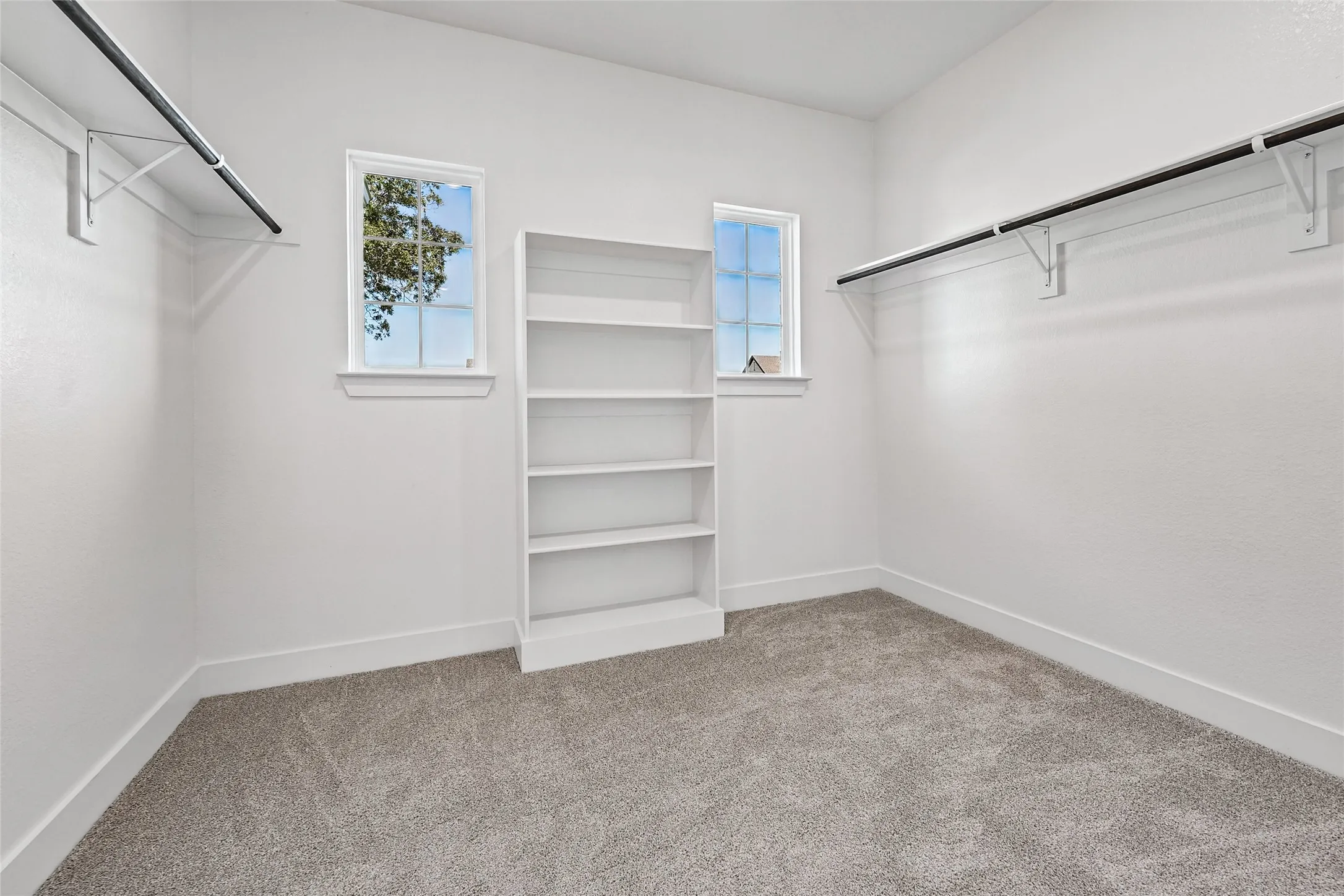 White walls with a textured finish, built-in shelving unit, dual windows with white trim, gray carpeting, and hanging rods with white brackets