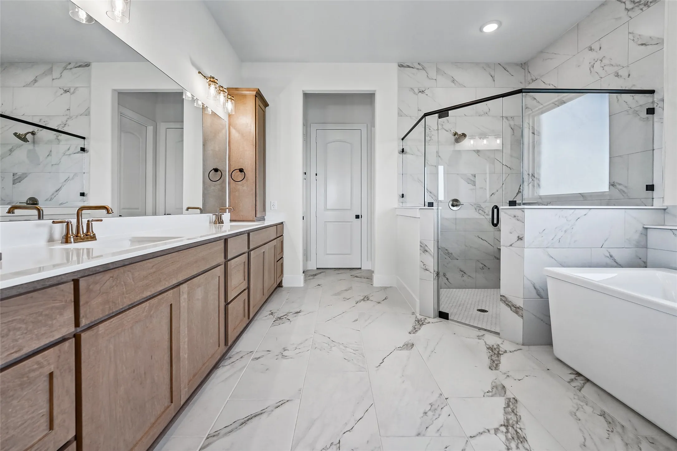 White marble-look floor and wall tile, dual vanity with wood-finish cabinetry, frameless glass shower enclosure, and a freestanding soaking tub