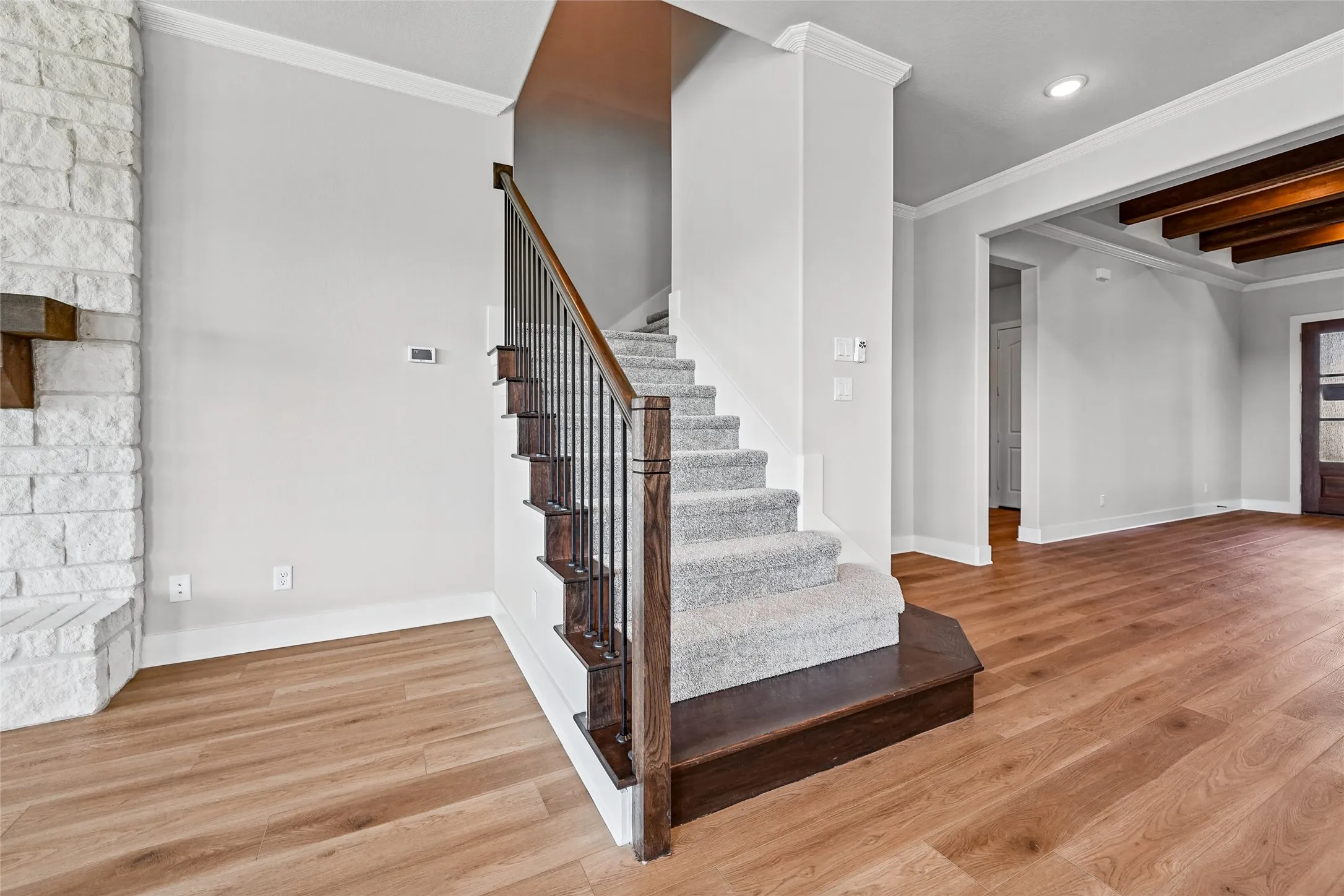 Light wood-finish flooring, a carpeted staircase with dark wood and iron railing, a stone-clad fireplace, crown molding, and exposed wood beams