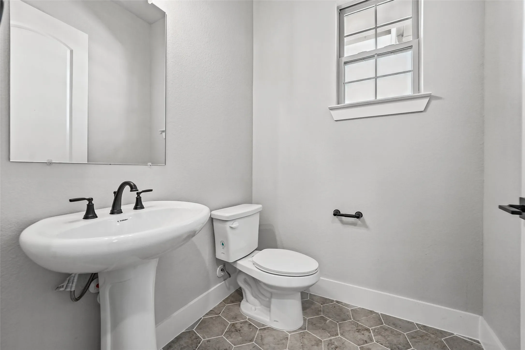 Pedestal sink with matte black faucet, hexagonal floor tiles, white baseboards, and a framed window