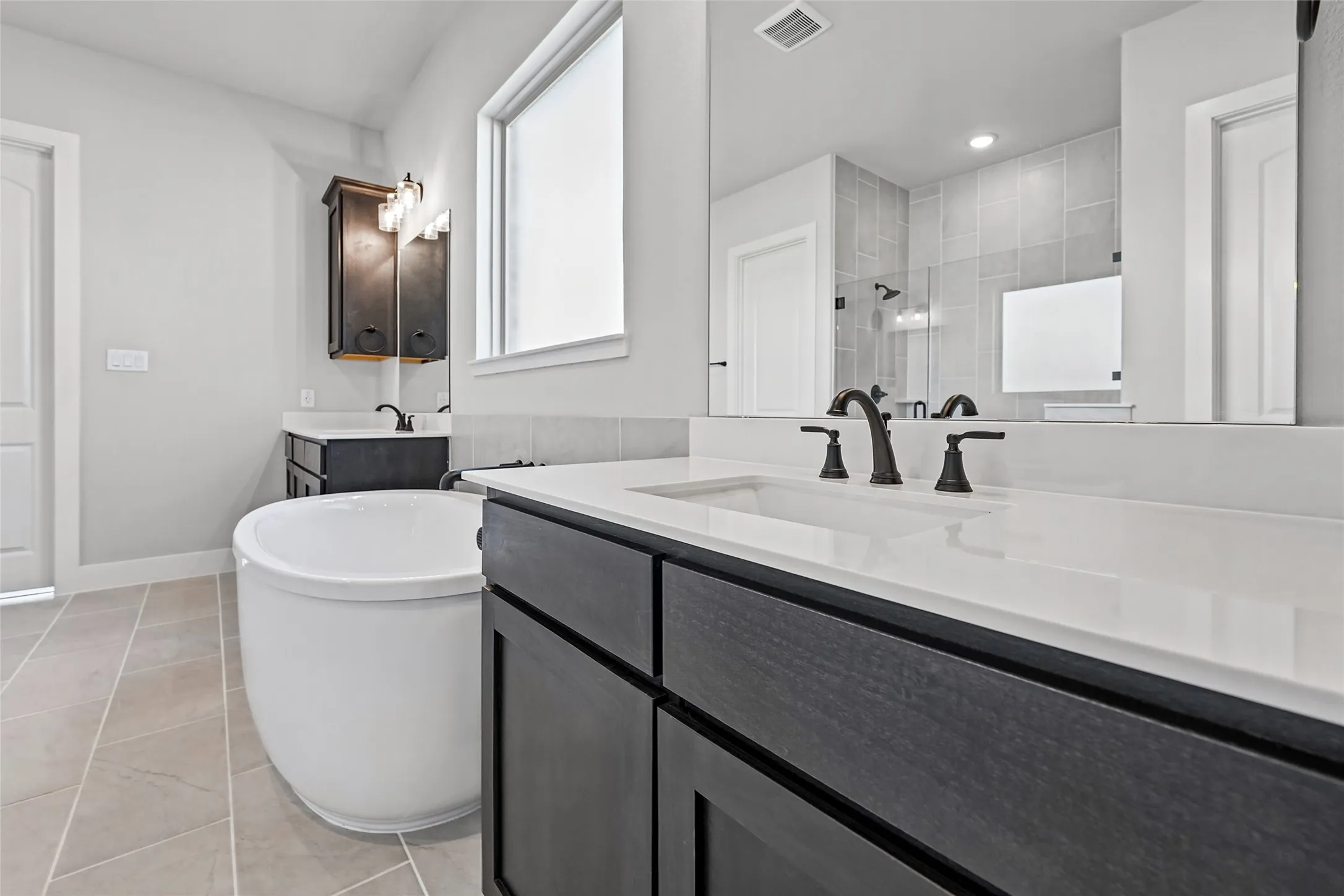 Freestanding soaking tub, dual dark wood-finish vanities with white countertops, matte black plumbing fixtures, and neutral tile flooring