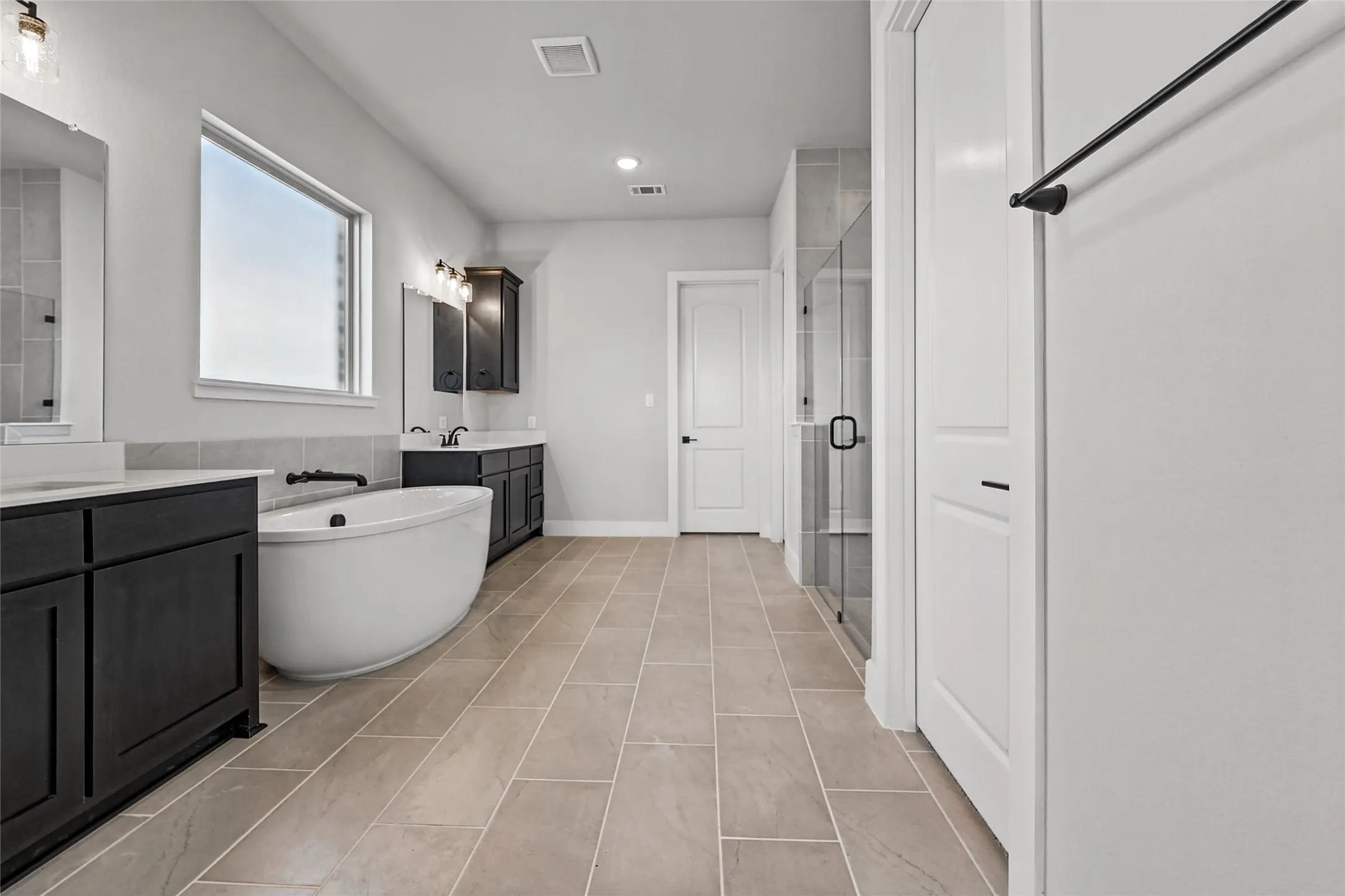 Dual vanity sinks, free-standing soaking tub, glass-enclosed shower, rectangular floor tiles, and a white interior door with black hardware