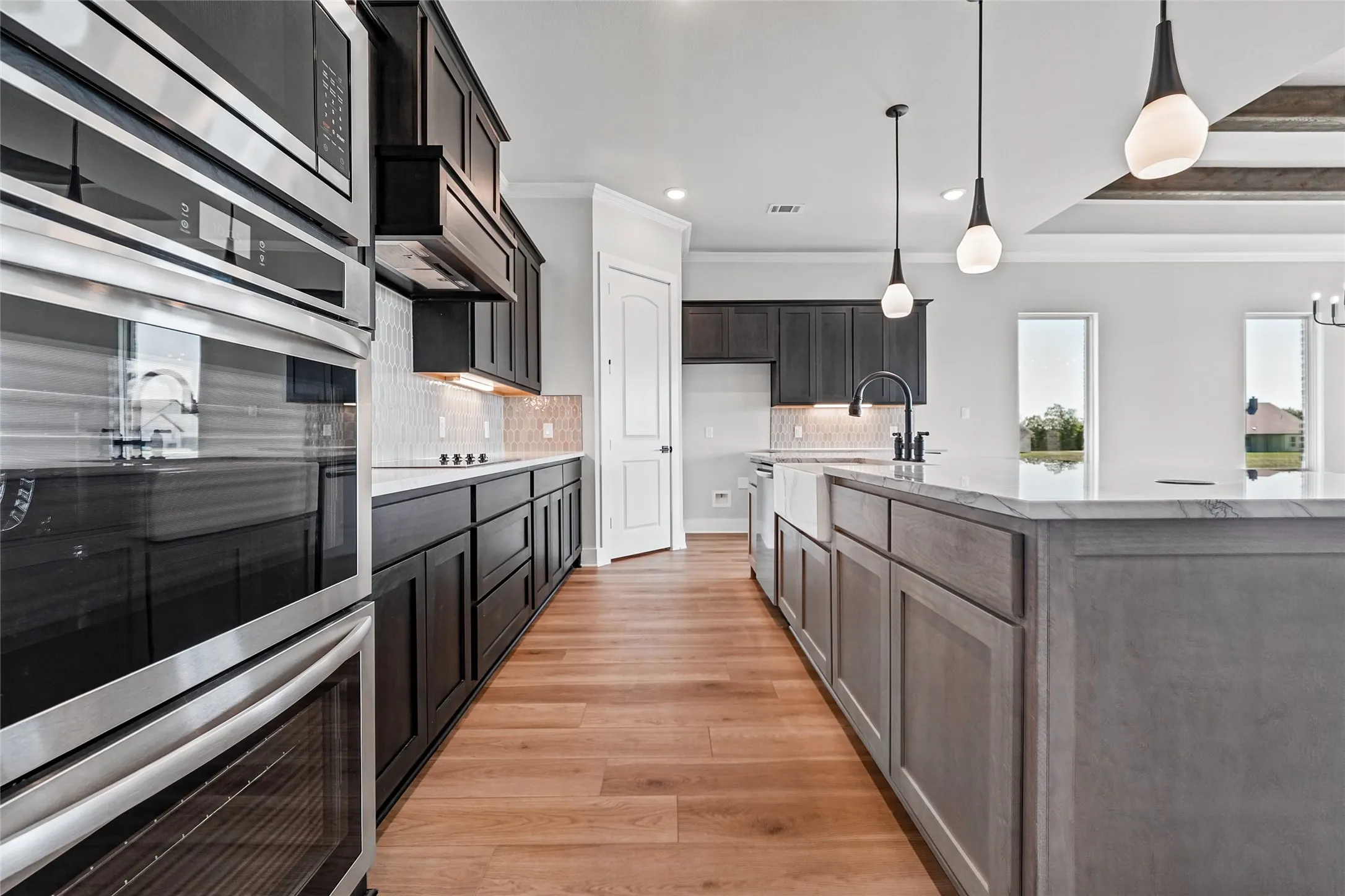 Stainless steel double oven and microwave, dark wood-finish cabinetry, light wood-finish flooring, white countertop surfaces, and an integrated farmhouse sink
