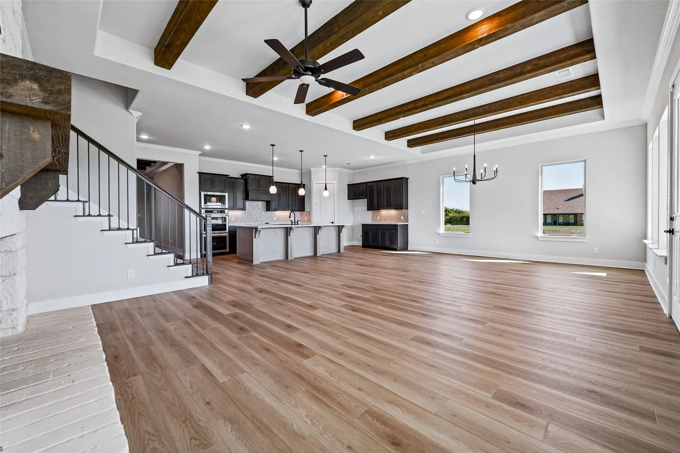 High ceilings with exposed wood beams, wood-finish flooring, a kitchen with dark cabinetry and stainless steel appliances, a staircase with metal balusters, and pendant lighting
