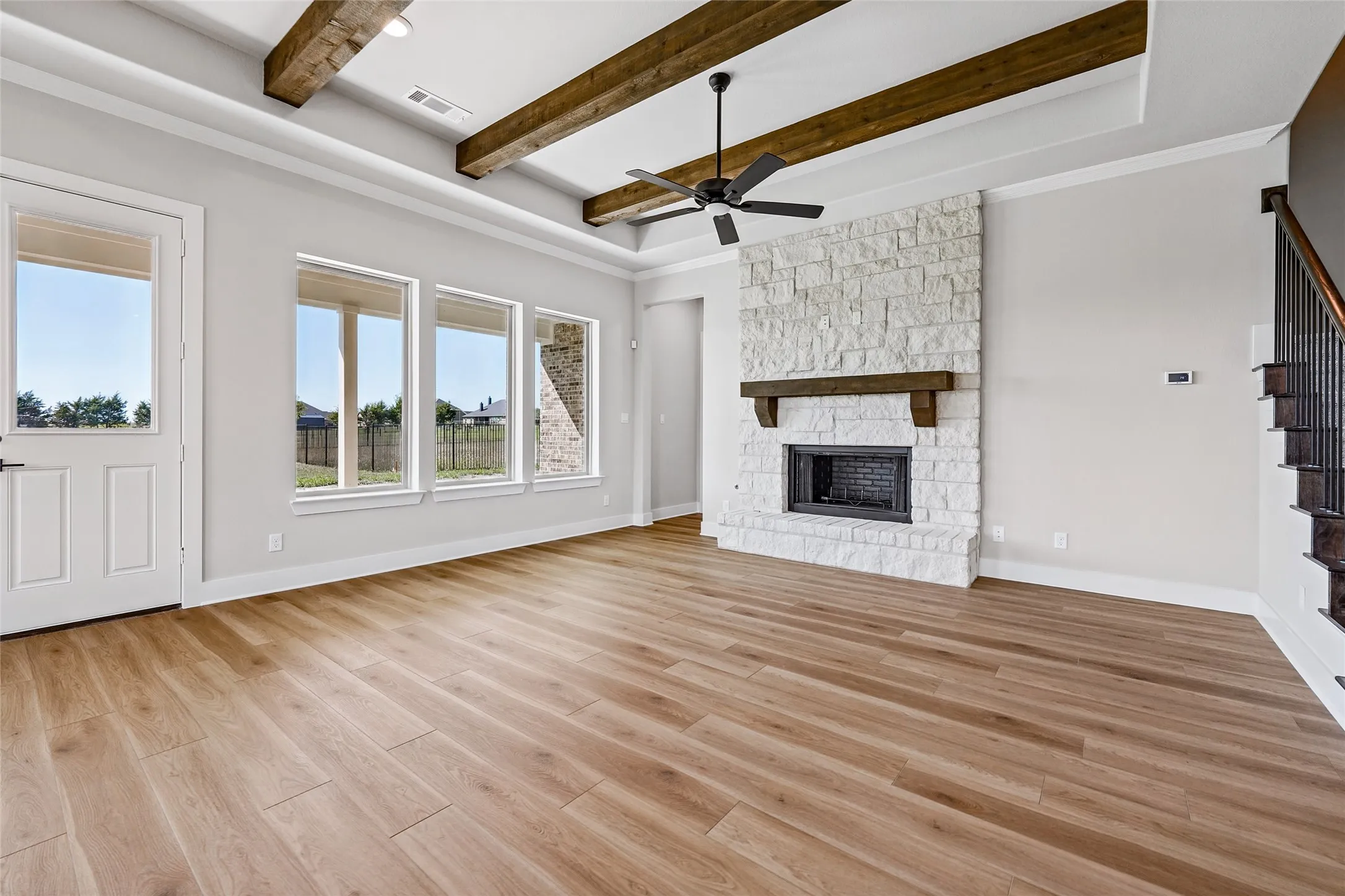 Wood-finish flooring, white stone fireplace with a dark wood mantel, exposed ceiling beams, ceiling fan, and multiple large windows